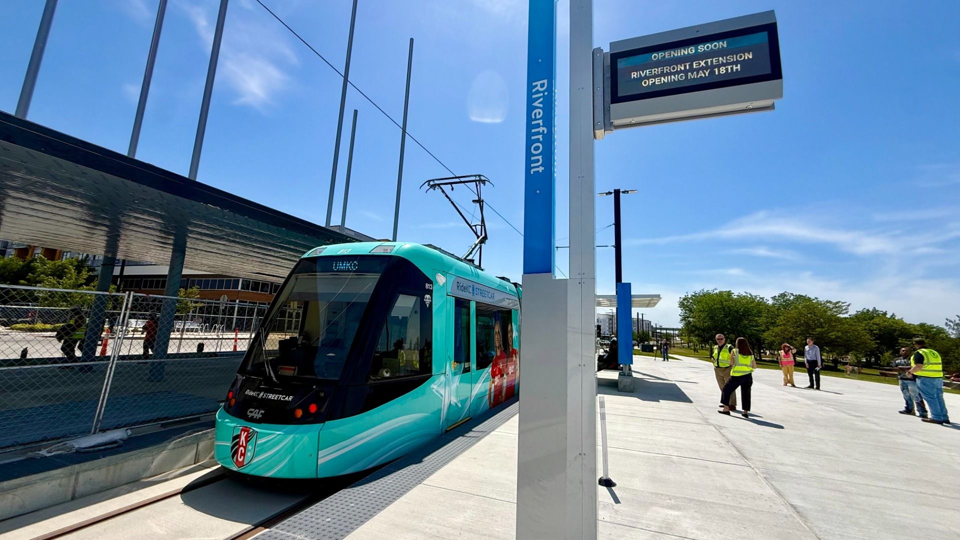 Blue-green modern streetcar at a riverfront station with overhead wires, a tall blue sign, a digital board reading "OPENING SOON", and workers in safety vests on a sunny day.