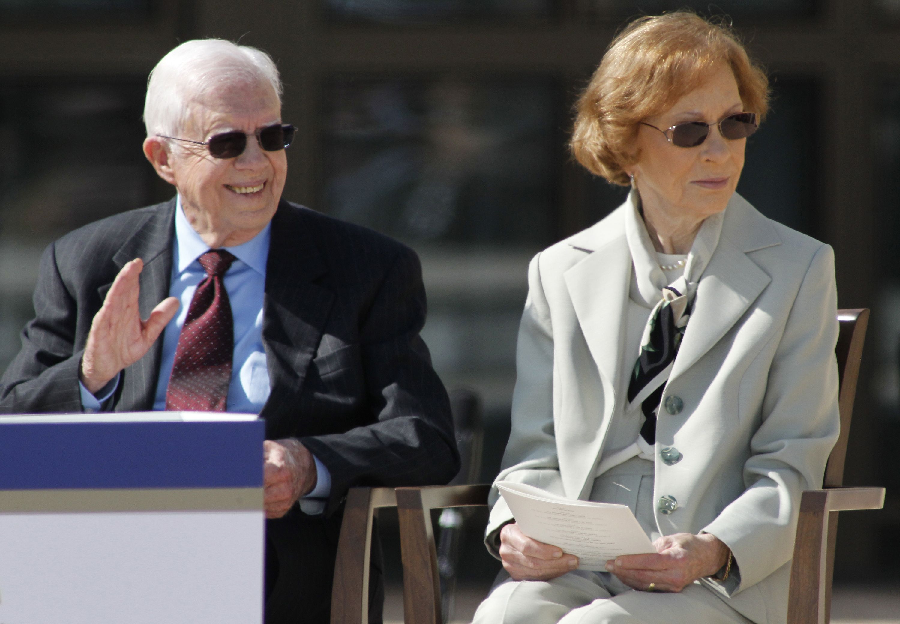 Jimmy Carter in a blue suit with a maroon tie and sunglasses, sitting next to wife Rosalynn in a gray suit