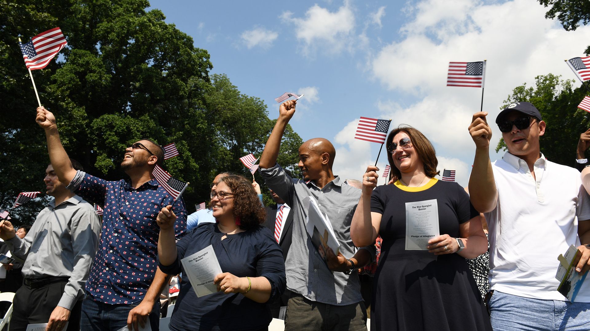 a group of people smiling while holding mini american flags. A blue sky with some clouds is behind them