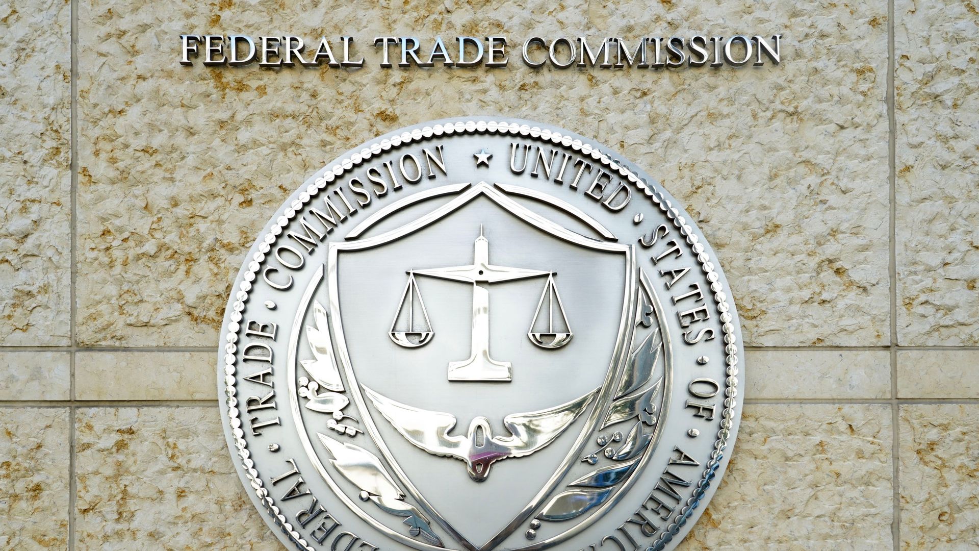 Close-up of the Federal Trade Commission sign on a beige stone wall. A large silver circular seal with scales of justice on a shield dominates, with the words "FEDERAL TRADE COMMISSION" above.