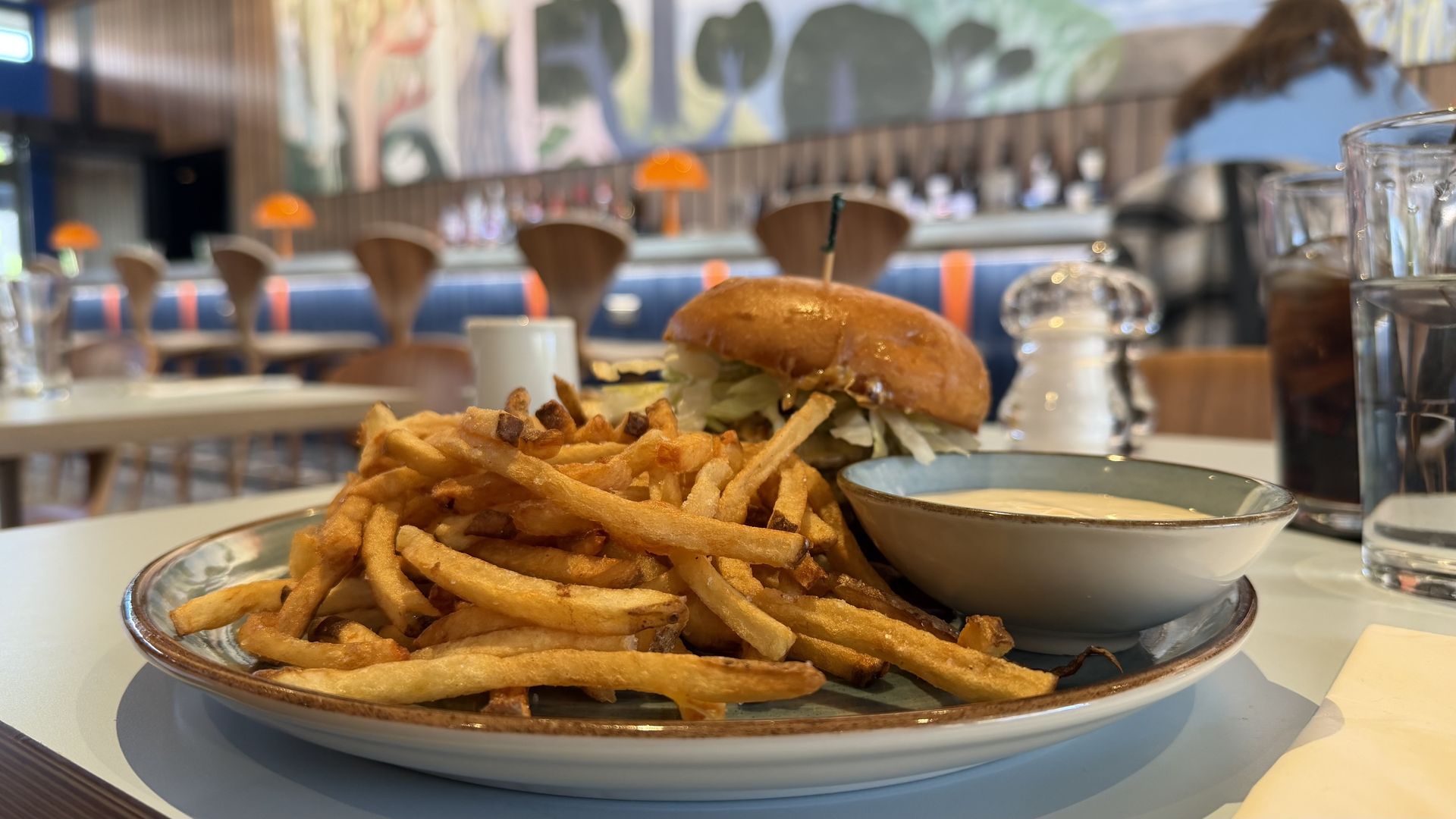 Burger with lettuce and a mound of french fries on a plate, in a bright casual restaurant with a blurred colorful mural and orange pendant lights in the background.