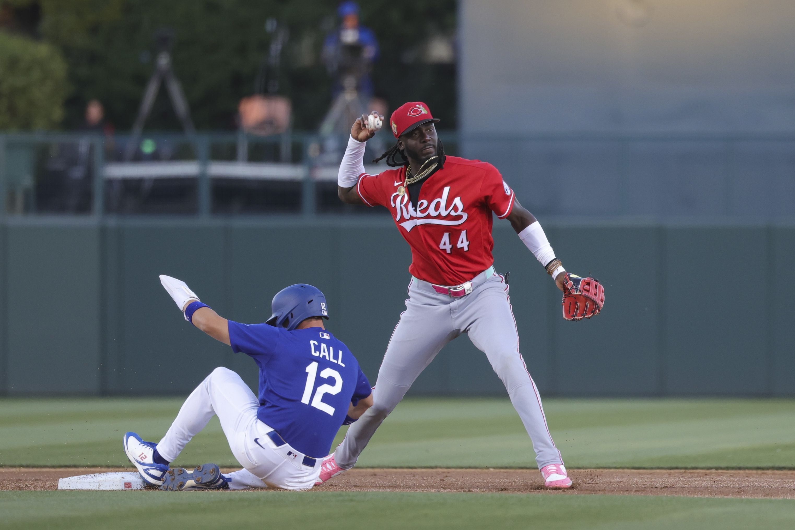 Reds player Elly De La Cruz, in a red jersey #44 and gray pants, throws from the infield while a blue-uniform baserunner slides into a base on the dirt.