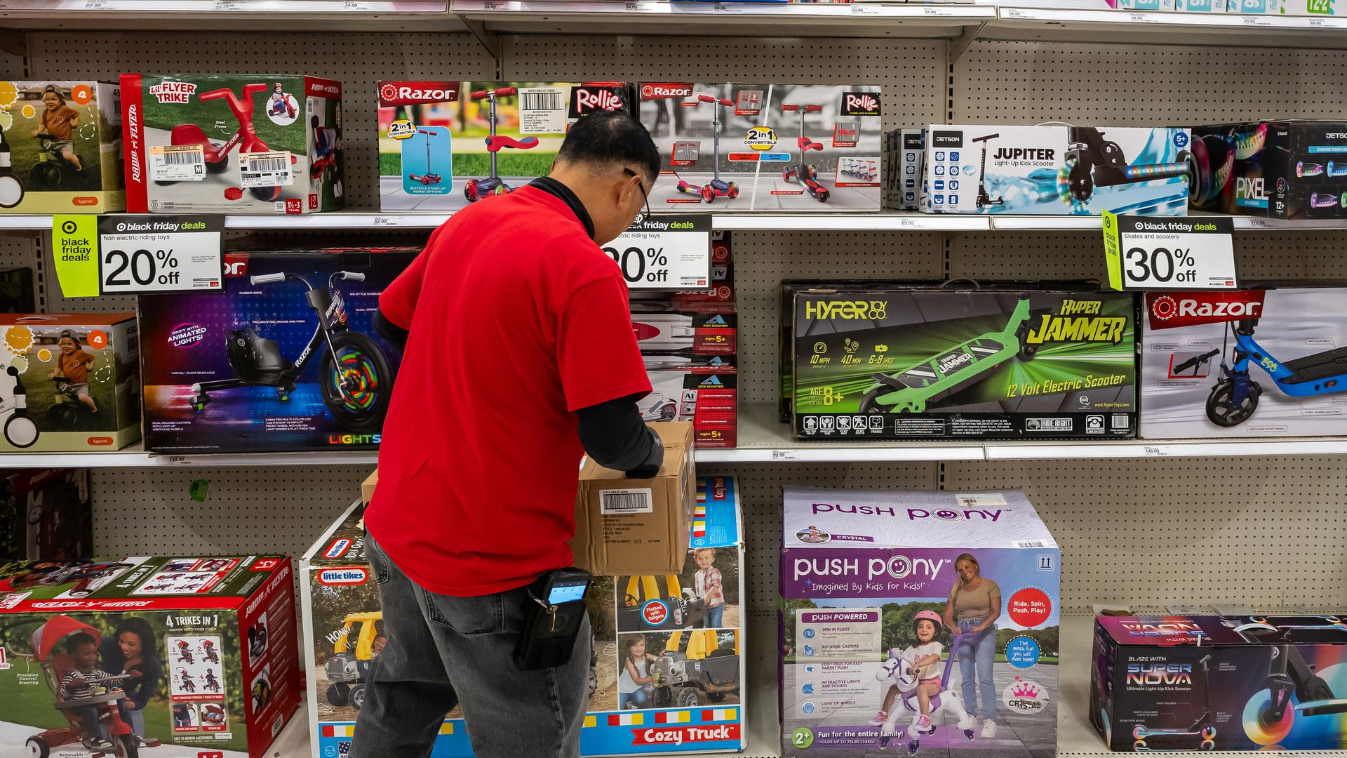A man in a bright red shirt browses a toy aisle, inspecting ride-on toys and scooters on metal shelves filled with colorful boxed packaging.