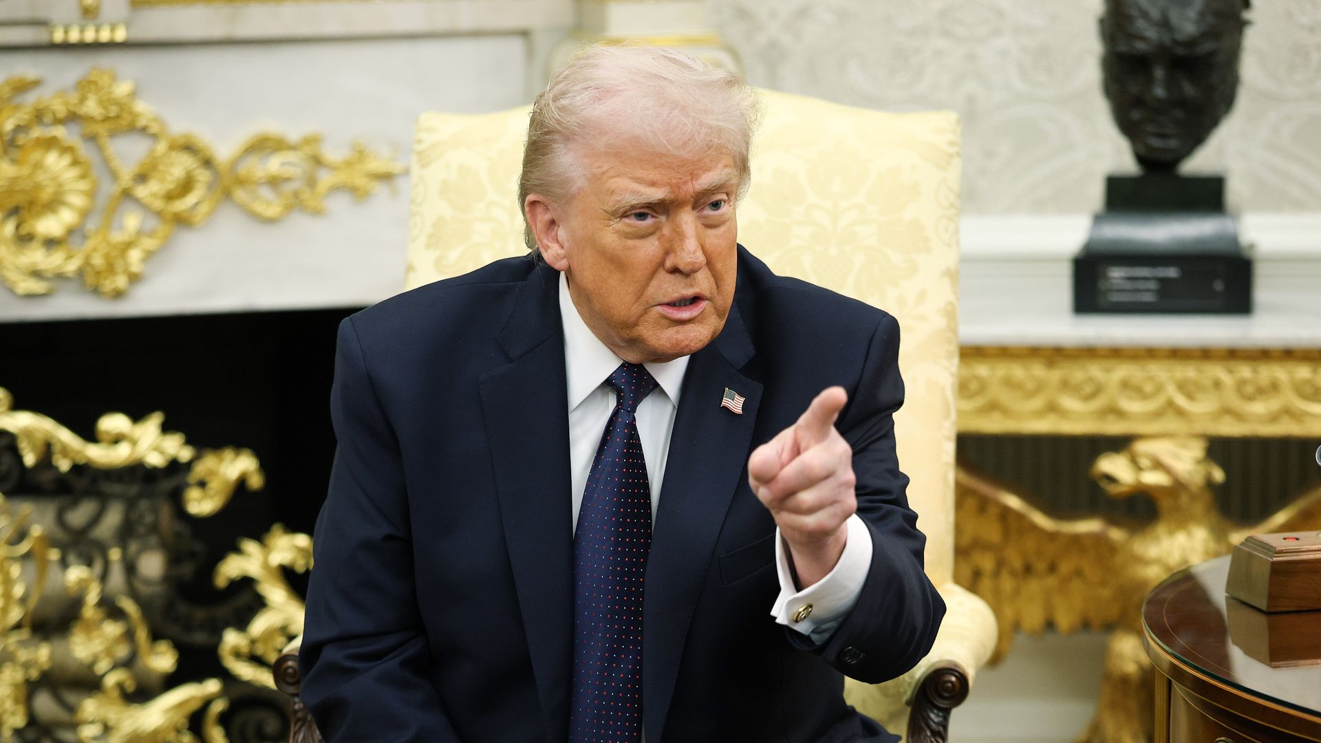 Trump sitting in a yellow chair with his index finger pointing, wearing a navy suit, white collared shirt and blue tie