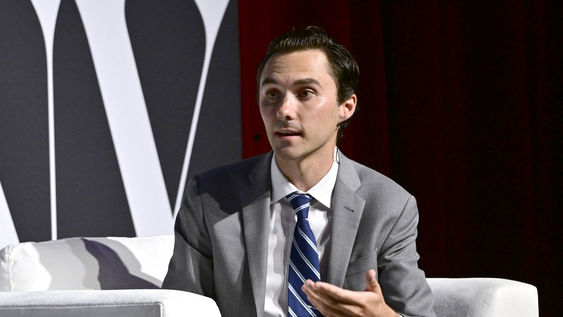 David Hogg in a gray suit, white shirt and blue and white striped tie speaks while seated in a white chair.