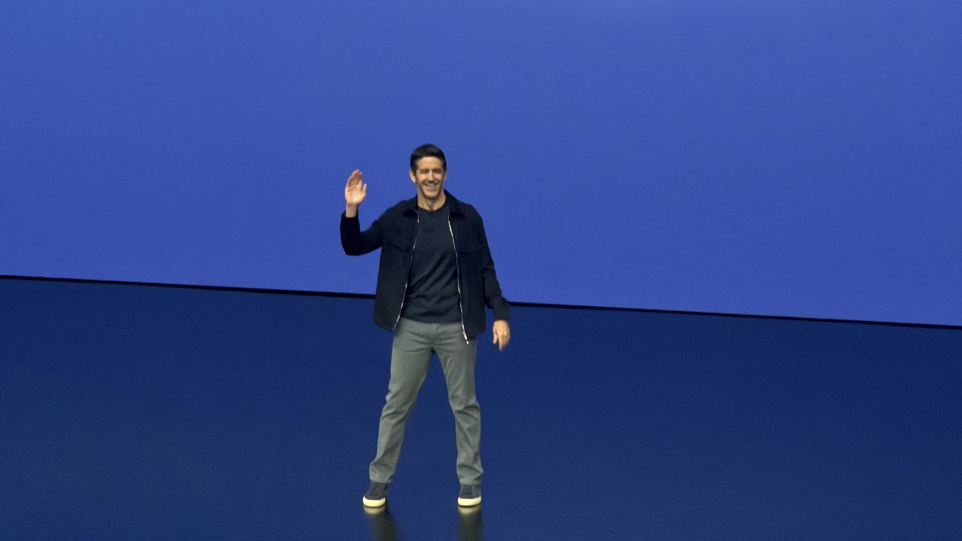 Man stands on a blue stage, waving with his right hand. He wears a dark jacket, black shirt, gray pants, and sneakers against a blue backdrop with a curved pink highlight.