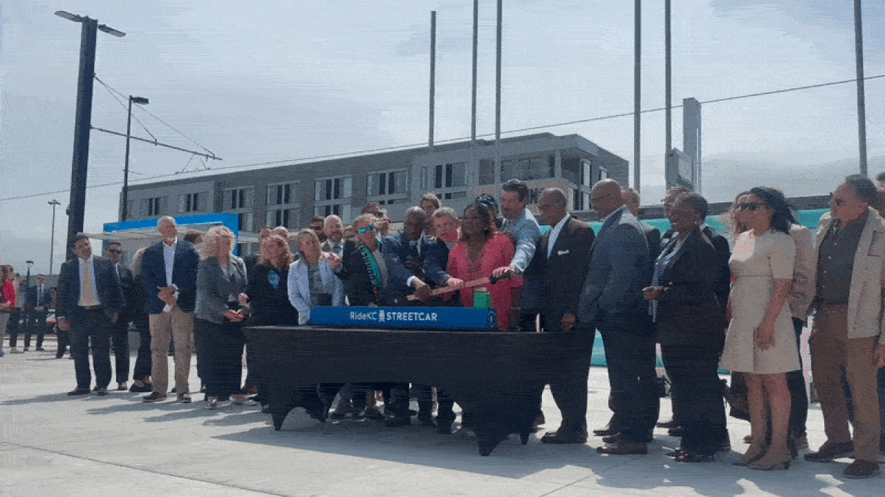 Diverse group of officials and professionals in business attire pose outdoors at a streetcar inauguration, pressing a ceremonial lever beside a blue RideKC Streetcar sign.