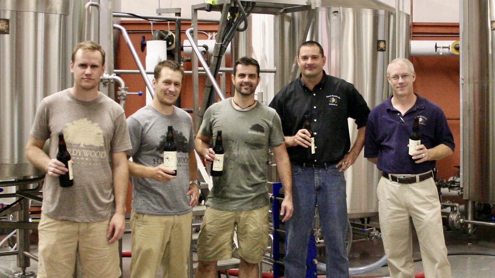 Five men stand in a brewery production room, each holding beer bottles, with large stainless steel fermentation tanks and pipes in the background.