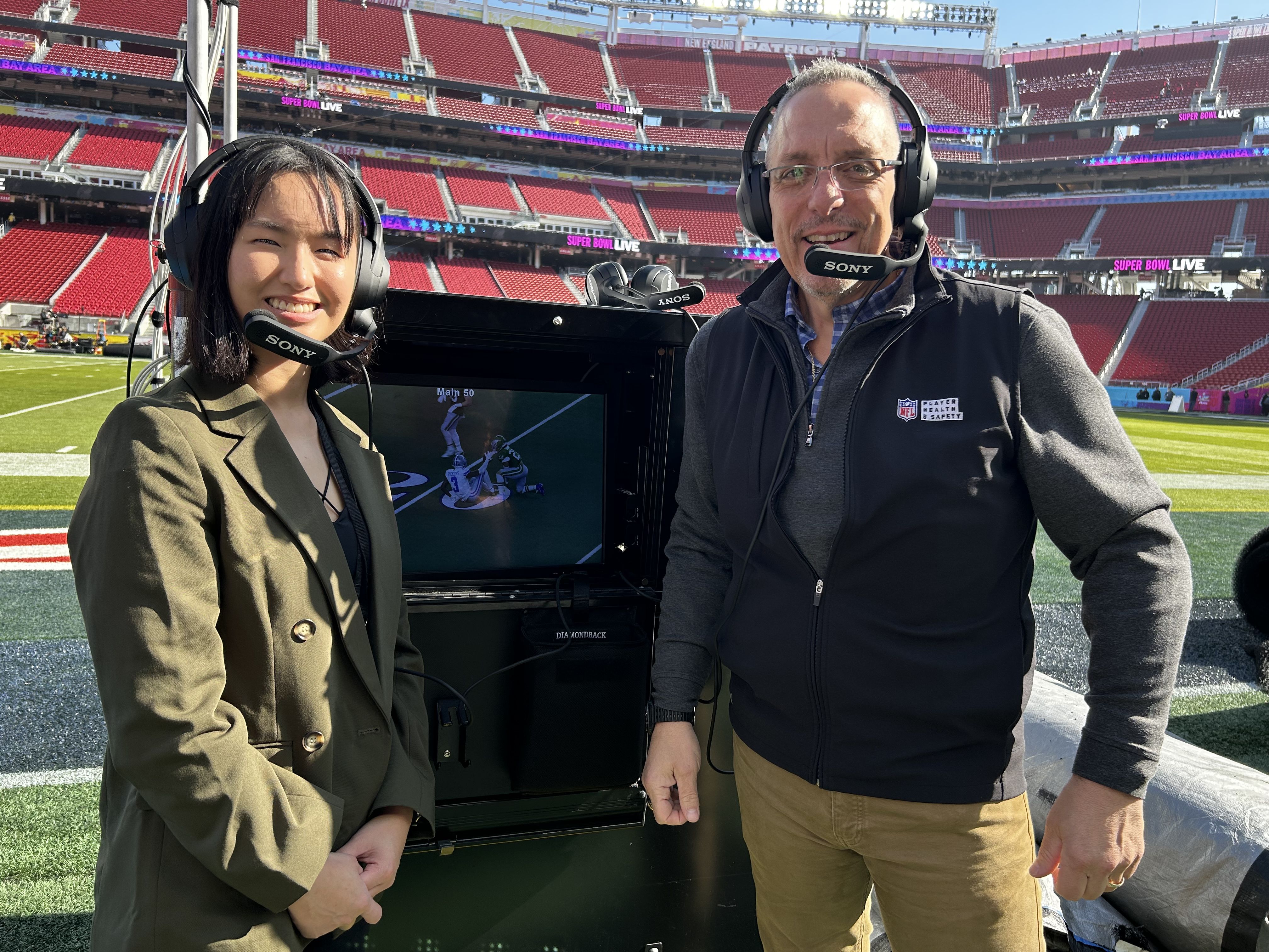 Two people wearing black Sony headset microphones stand on a football field near a monitor showing a play, with empty red stadium seats and "SUPER BOWL LIVE" banners in the background.