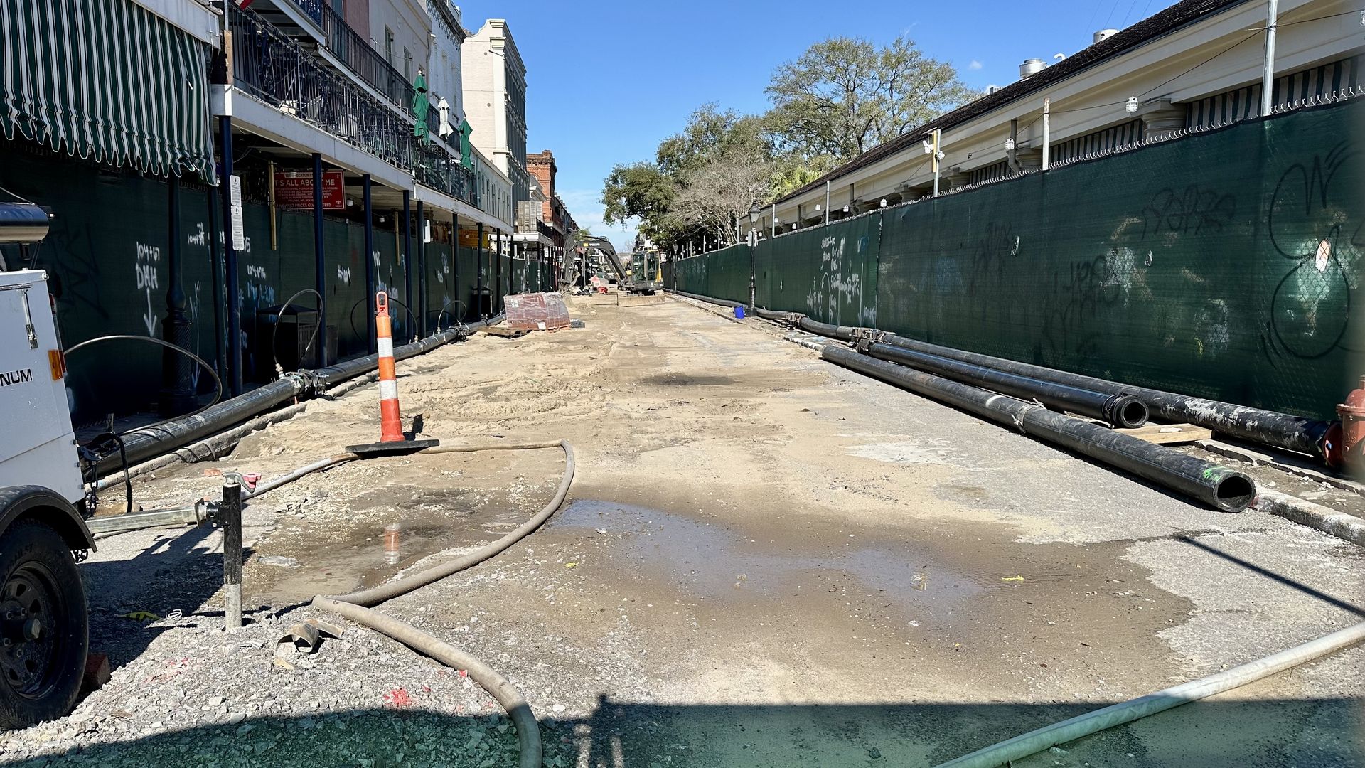 Construction site on a street with dirt and puddles, orange traffic cone, pipes along green fences on both sides, buildings on the left, clear blue sky overhead.