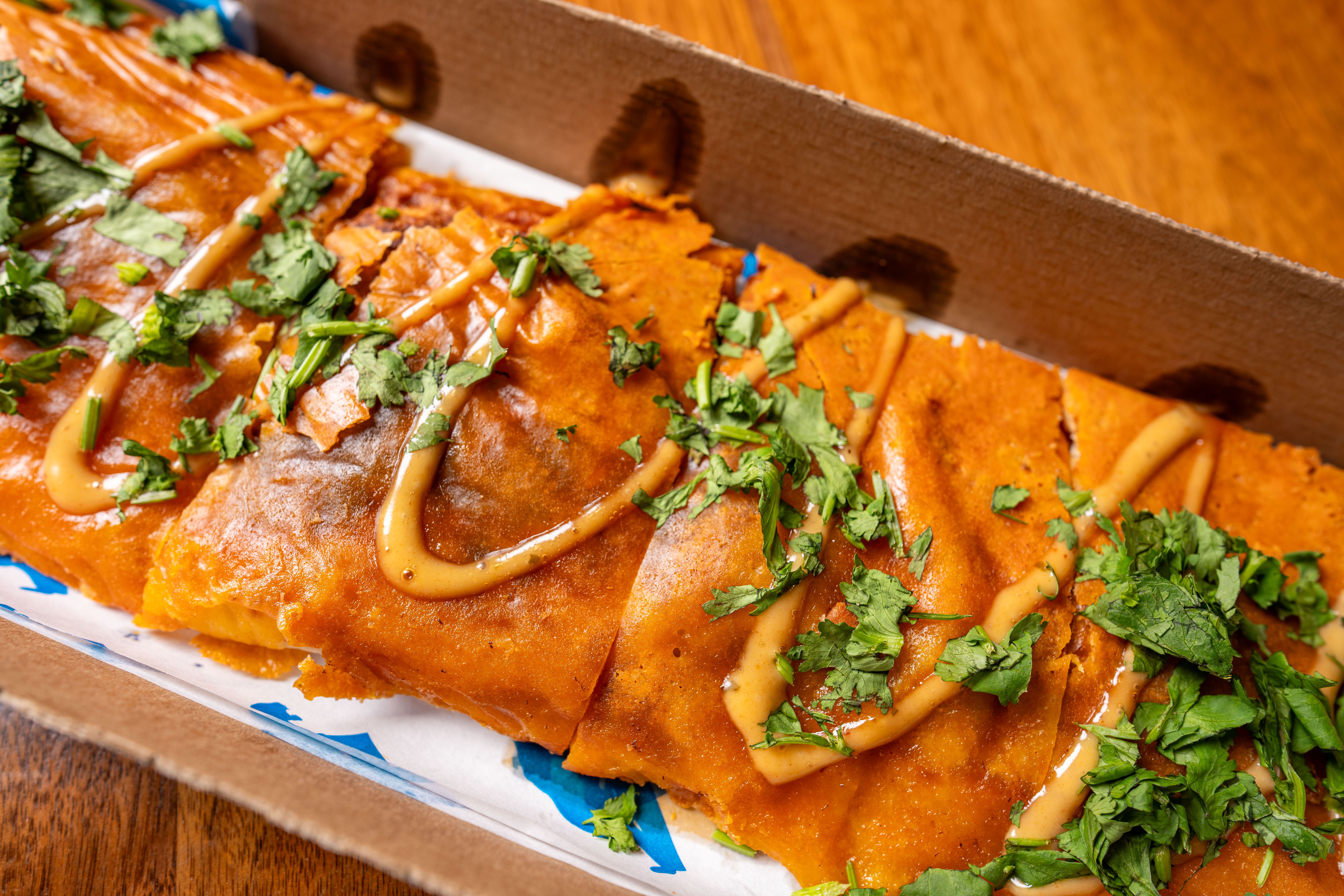 Close-up of orange fried tortillas (tostadas) in a cardboard takeout box, topped with chopped cilantro and a light drizzle of sauce on a wooden table.