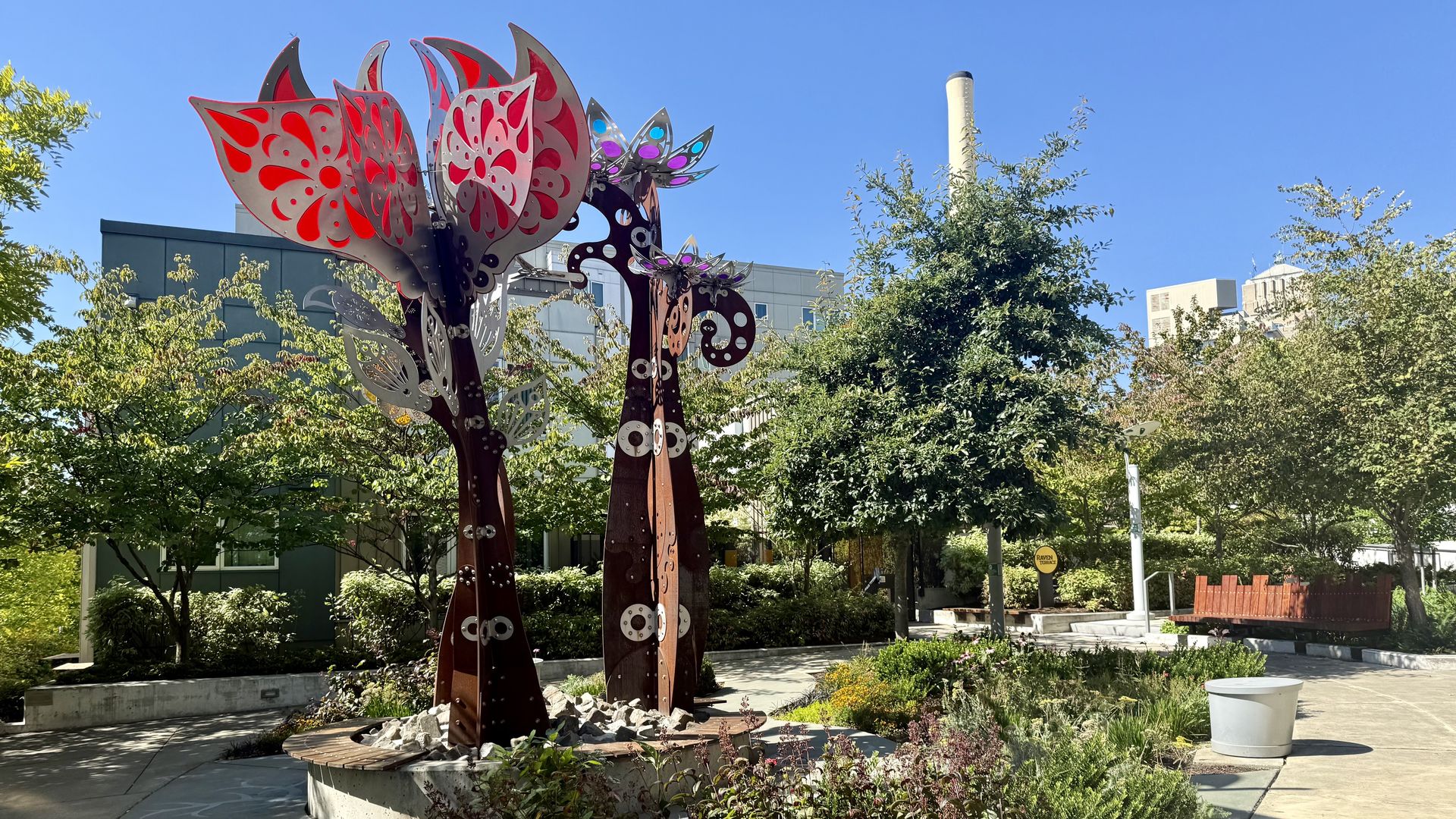 A view of a courtyard with a tall metal and colorful sculptures, with blue sky above and trees planted between buildings.