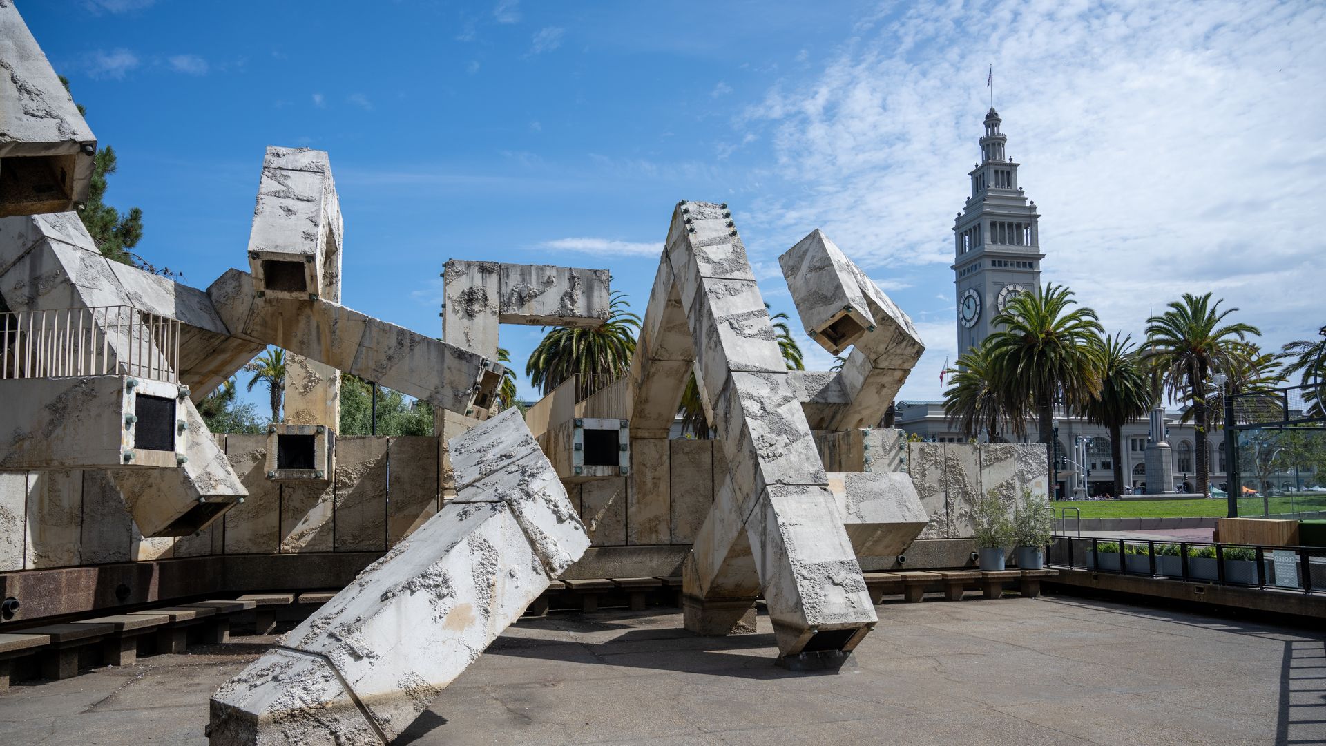 Large, abstract gray concrete sculpture with geometric shapes in an open plaza, palm trees behind it, and a clock tower under a partly cloudy blue sky.
