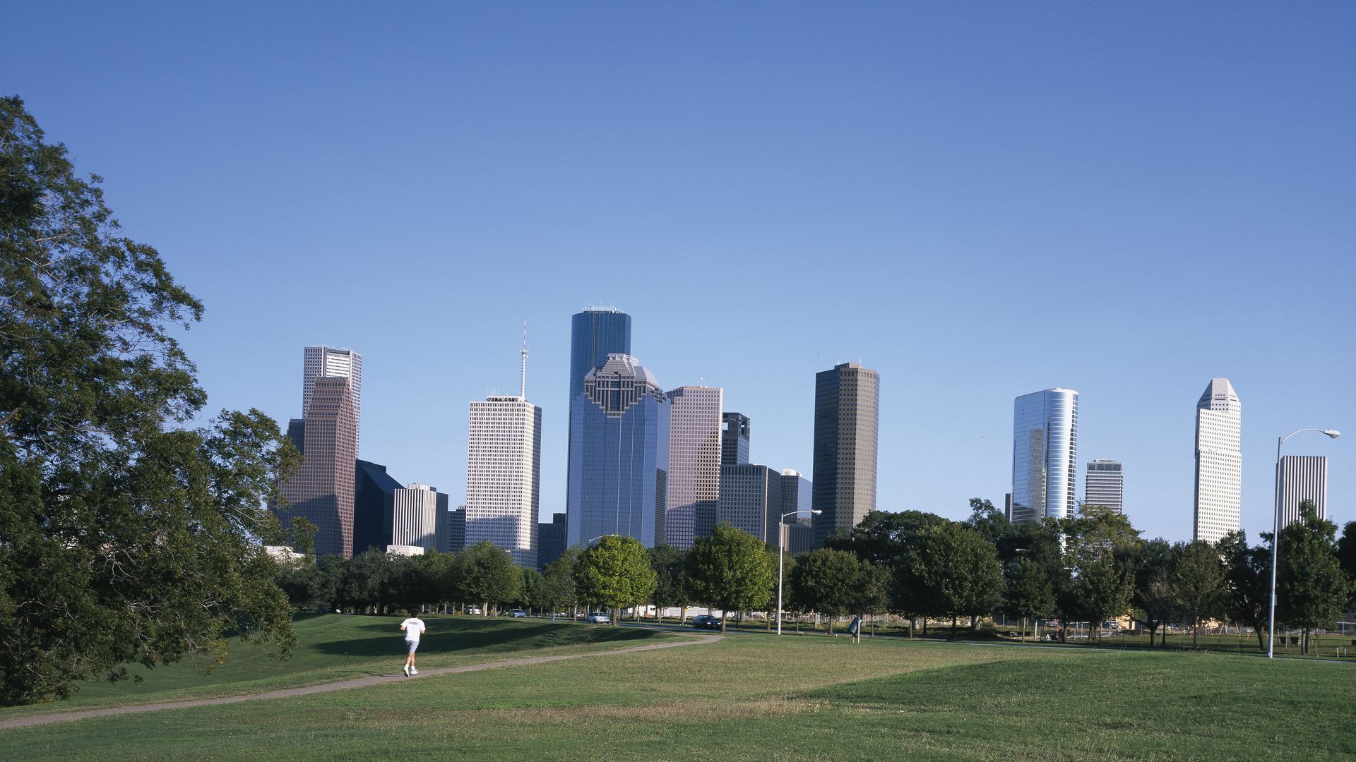 A person in the distance jogs up a path in a green meadow with the Houston skyline in the background