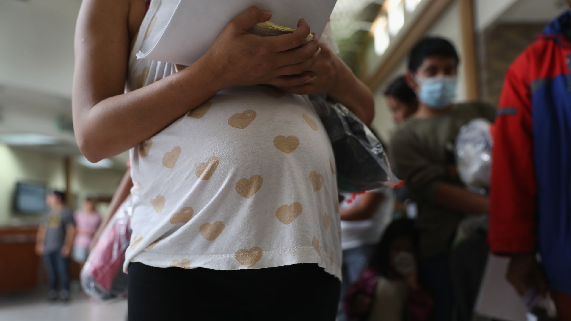 A pregnant woman holds papers and stands in line.