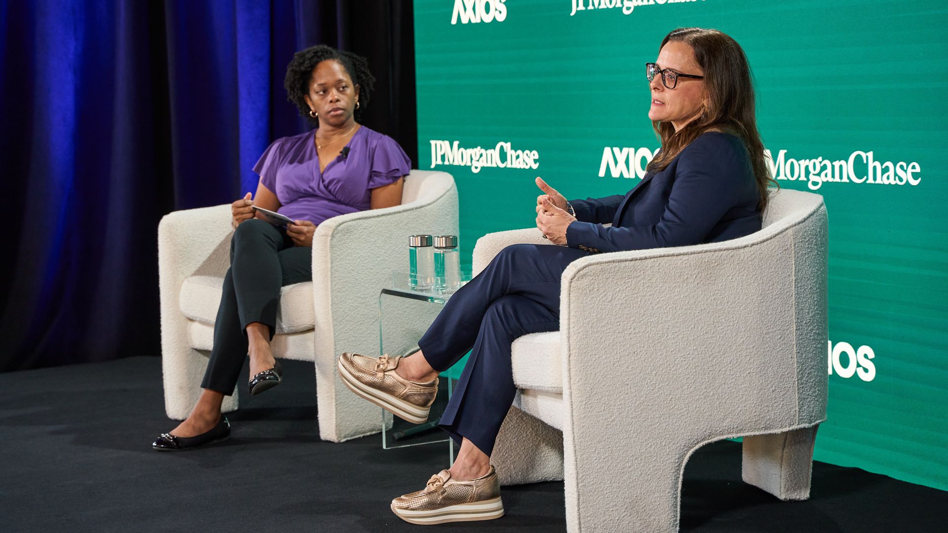 Two women seated on white armchairs on stage during a discussion, one in a purple blouse and black pants, the other in a navy suit and gold shoes, with JPMorgan Chase and AXIOS logos behind them.