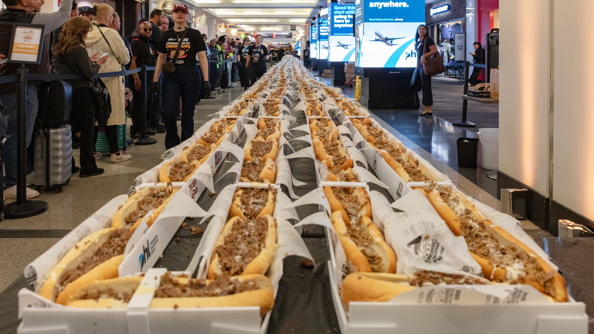 Inside a busy airport terminal, rows of long Philly cheesesteaks in white boxes along a central table, with crowds on both sides.