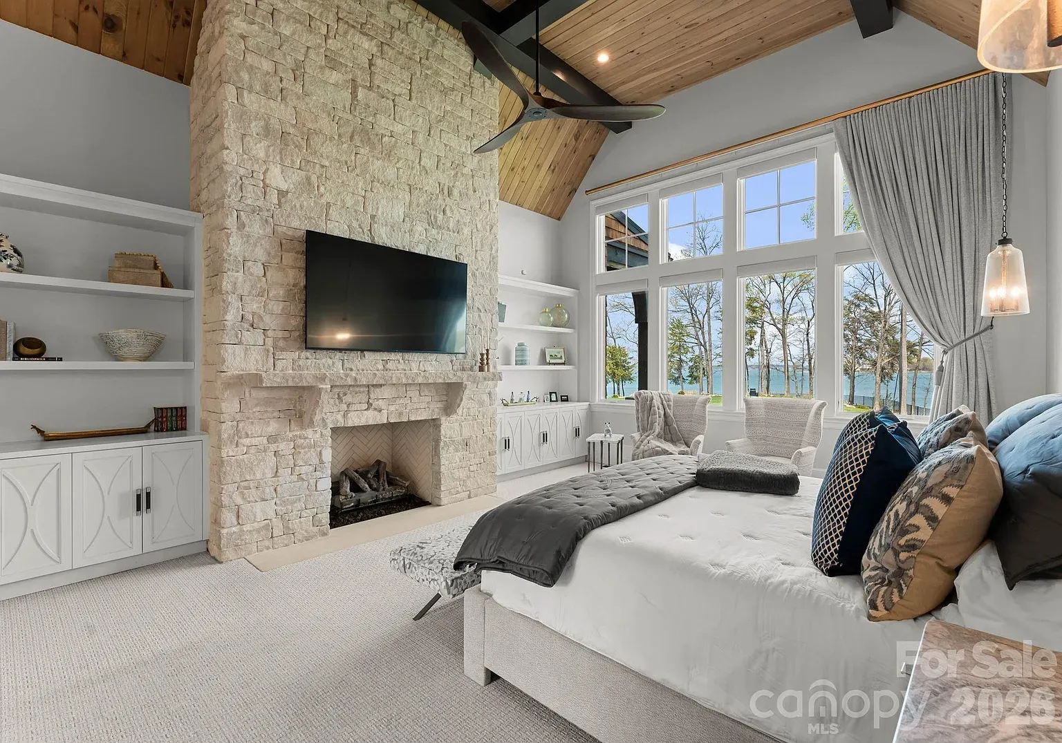 Bright bedroom featuring a tall stone fireplace with a mounted TV, white built-in shelves, and large windows with gray curtains overlooking the ocean. A plush bed sits opposite.