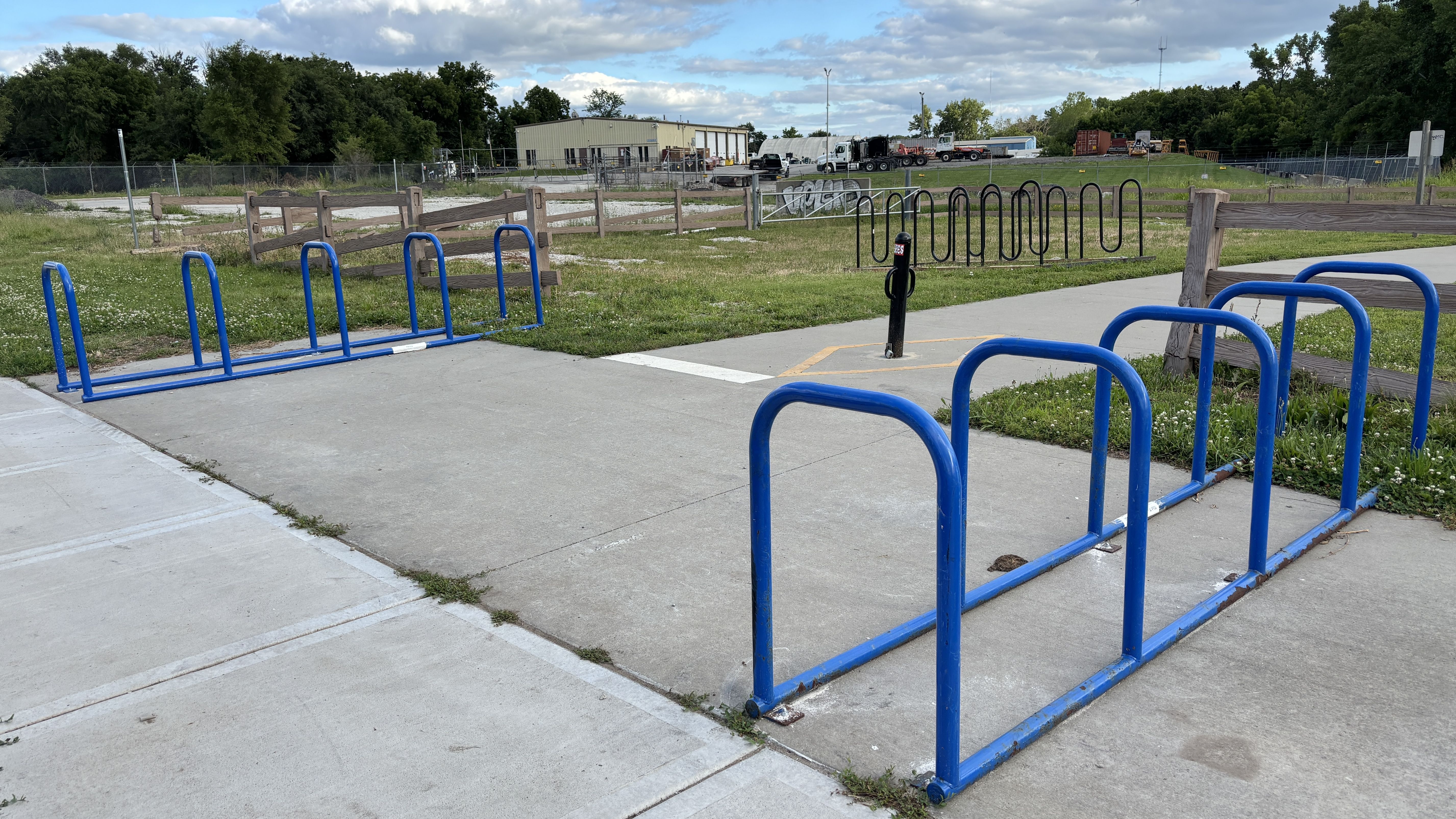 Wide park scene with bright blue bike racks in the foreground, a wooden fence, grassy field, and distant industrial buildings and trucks under a partly cloudy sky.