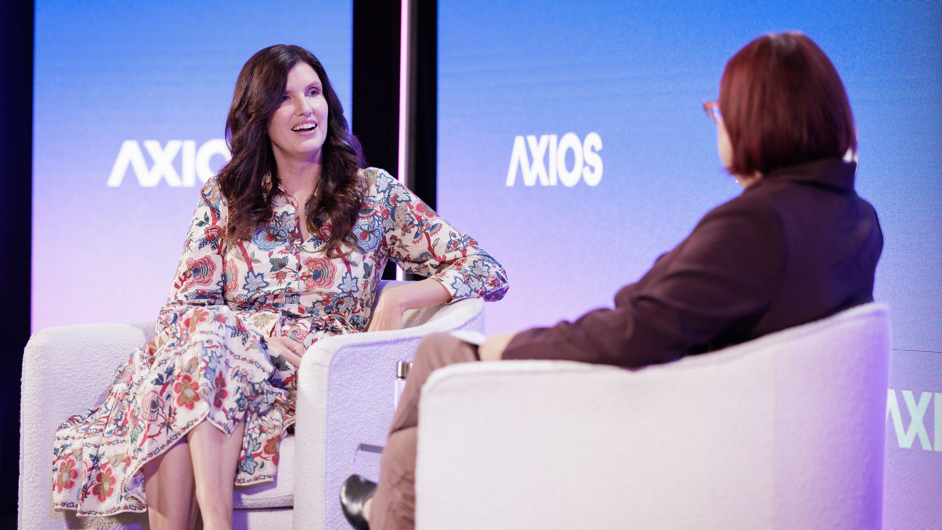 Two people seated in white armchairs on a stage with blue backgrounds displaying the word AXIOS. One in a floral dress faces the camera, the other wears dark clothes with back to camera.