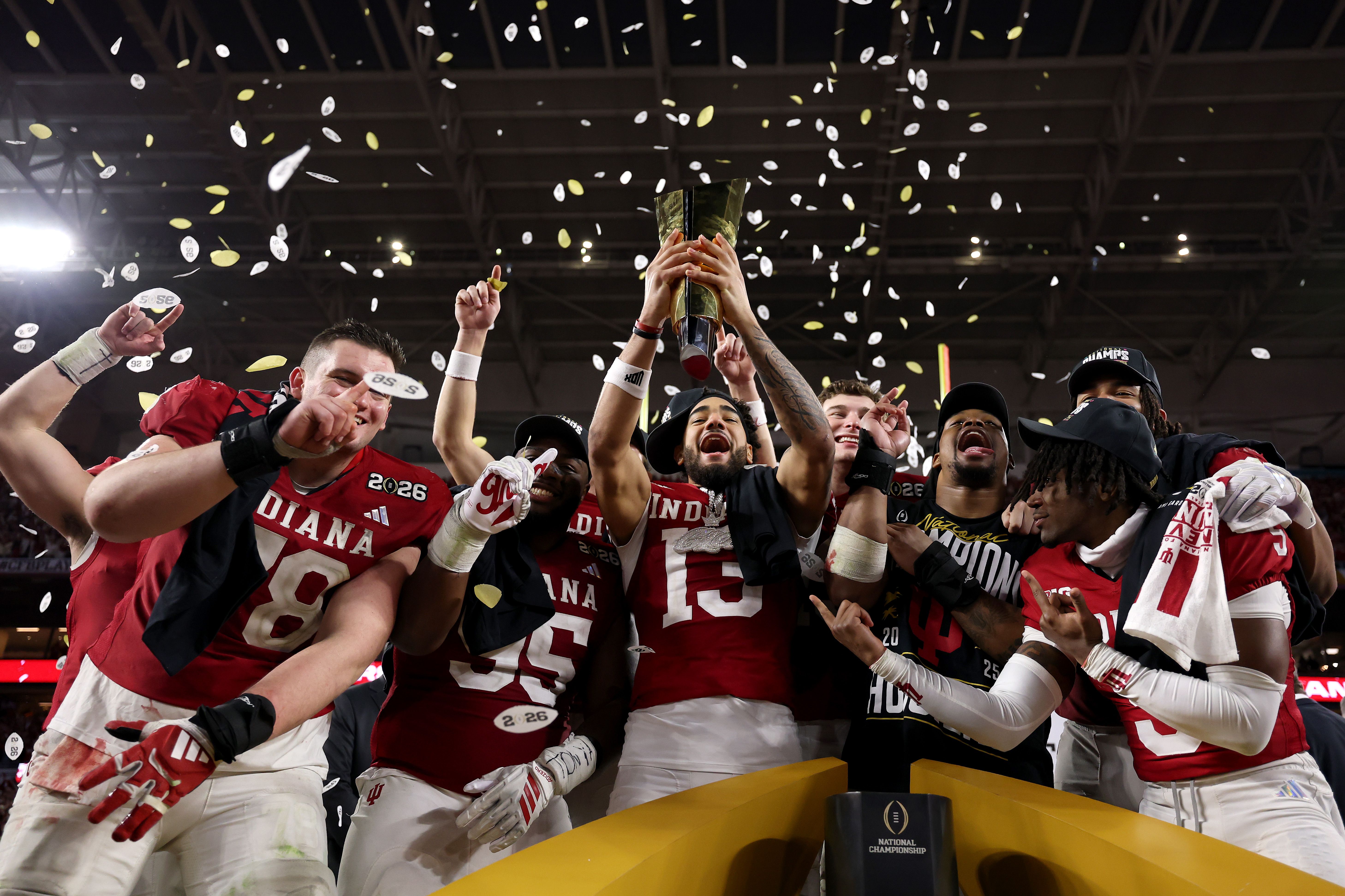 Indiana football players in red and white celebrate with a trophy amid falling confetti in a stadium, showing excitement and joy after a championship win.