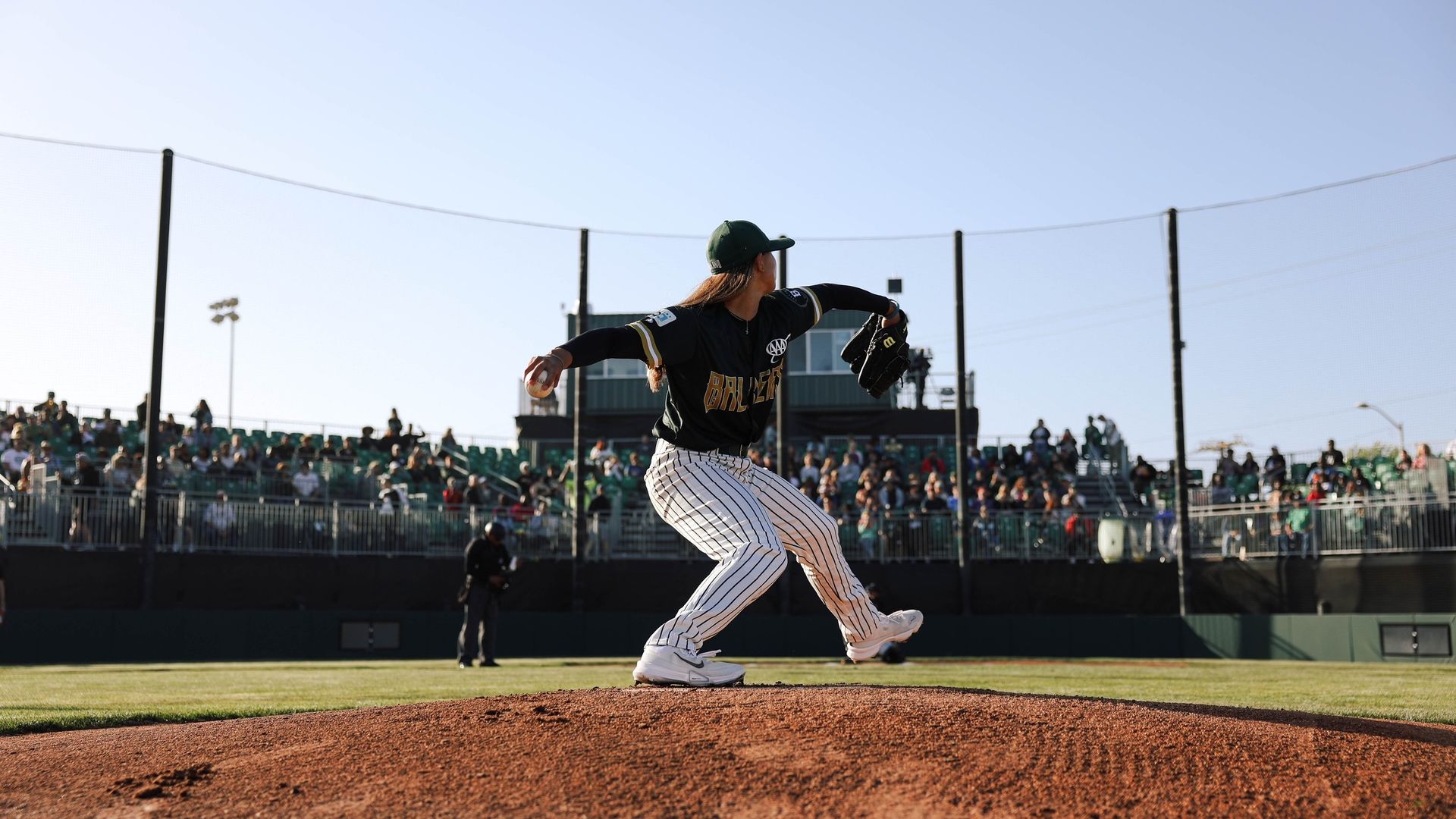 Baseball pitcher Kelsie Whitmore in black and yellow jersey with white striped pants winding up to throw on a mound with spectators and blue sky in the background.