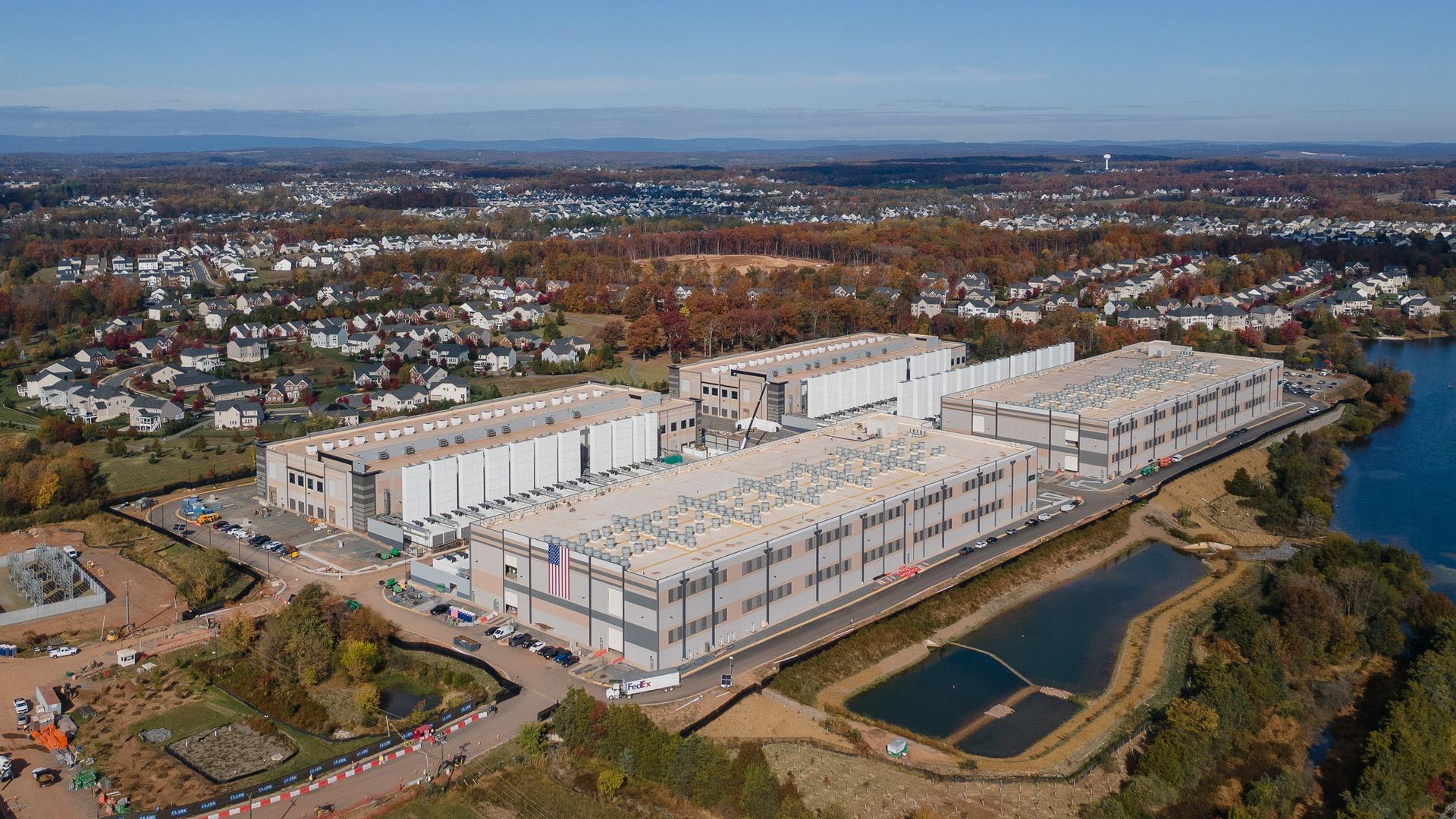 Aerial view of a large industrial complex with three beige and gray buildings featuring ventilation units on rooftops, an American flag, surrounded by a water body and suburban houses under a blue sky.