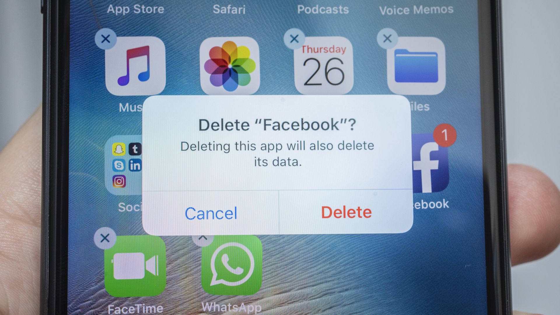 Close-up of an iPhone home screen showing a Delete "Facebook" confirmation dialog; behind it are app icons (Music, Photos, Calendar Thursday 26, Facebook) and a finger at the bottom.