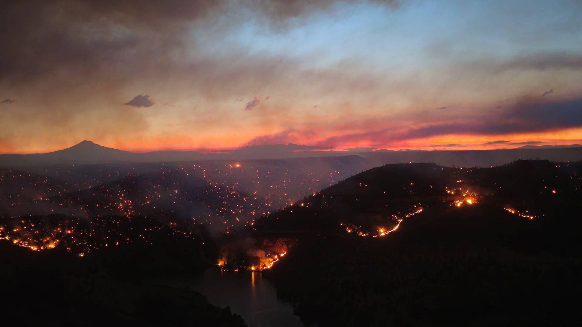 Wide sunset panorama with a distant volcano on the horizon, dark foreground hills, a smoky blue sky, and glowing orange embers scattered across the valley.