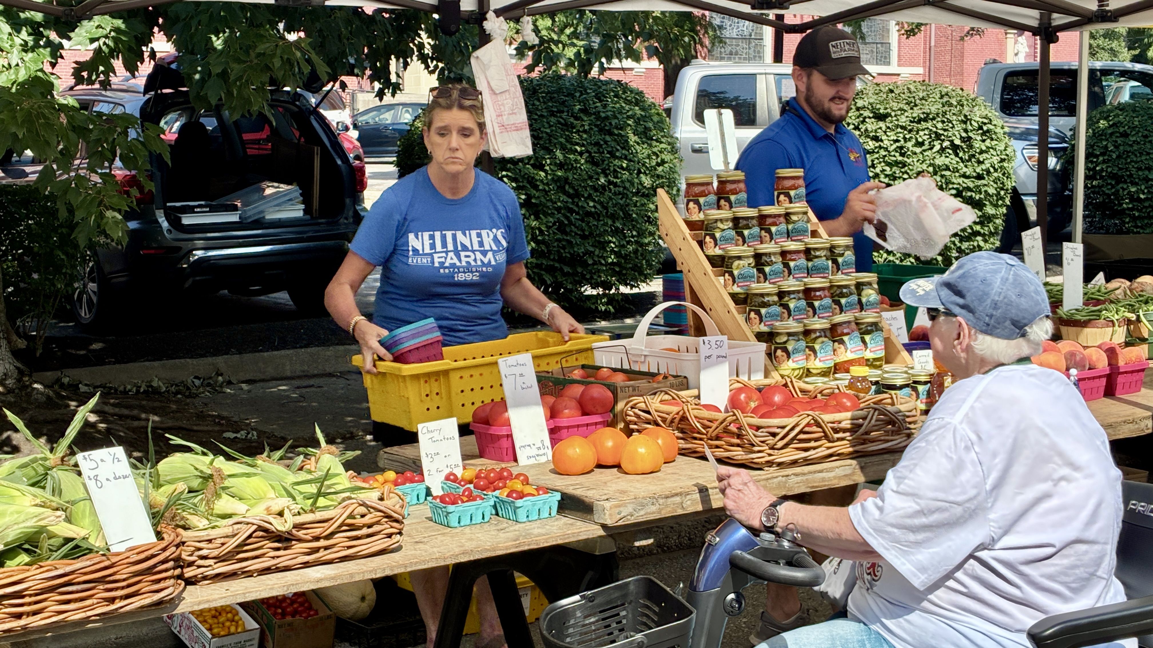 Outdoor market stall under a canopy. A woman in a blue Neltner's Farm shirt stacks produce; jars line the stand as a man in blue bags groceries, with a customer on a scooter nearby.