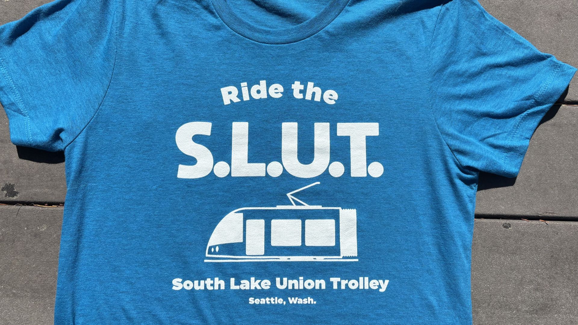 Blue T-shirt laid on a wooden surface, white text reads Ride the S.L.U.T. with a simple trolley illustration and the words South Lake Union Trolley Seattle, Wash.