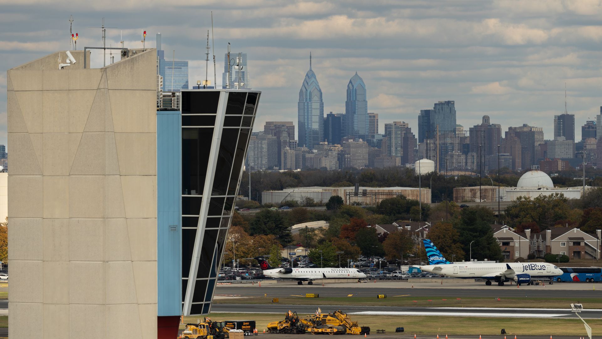 A JetBlue airplane wheels past an FAA air traffic control tower at Philadelphia International Airport with the city skyline in the background under mostly cloudy skies.