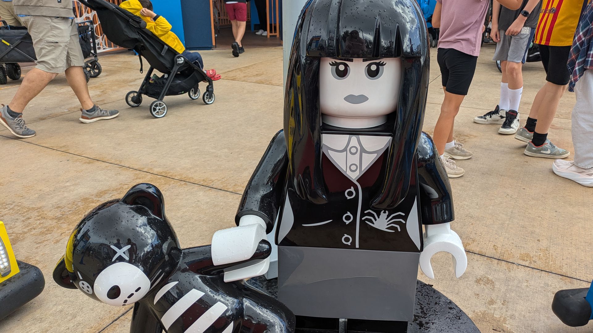 Tall glossy black LEGO-style statue of a woman with bangs in a suit stands outdoors, flanked by panda-faced motorcycles; people and a stroller are visible in the background.