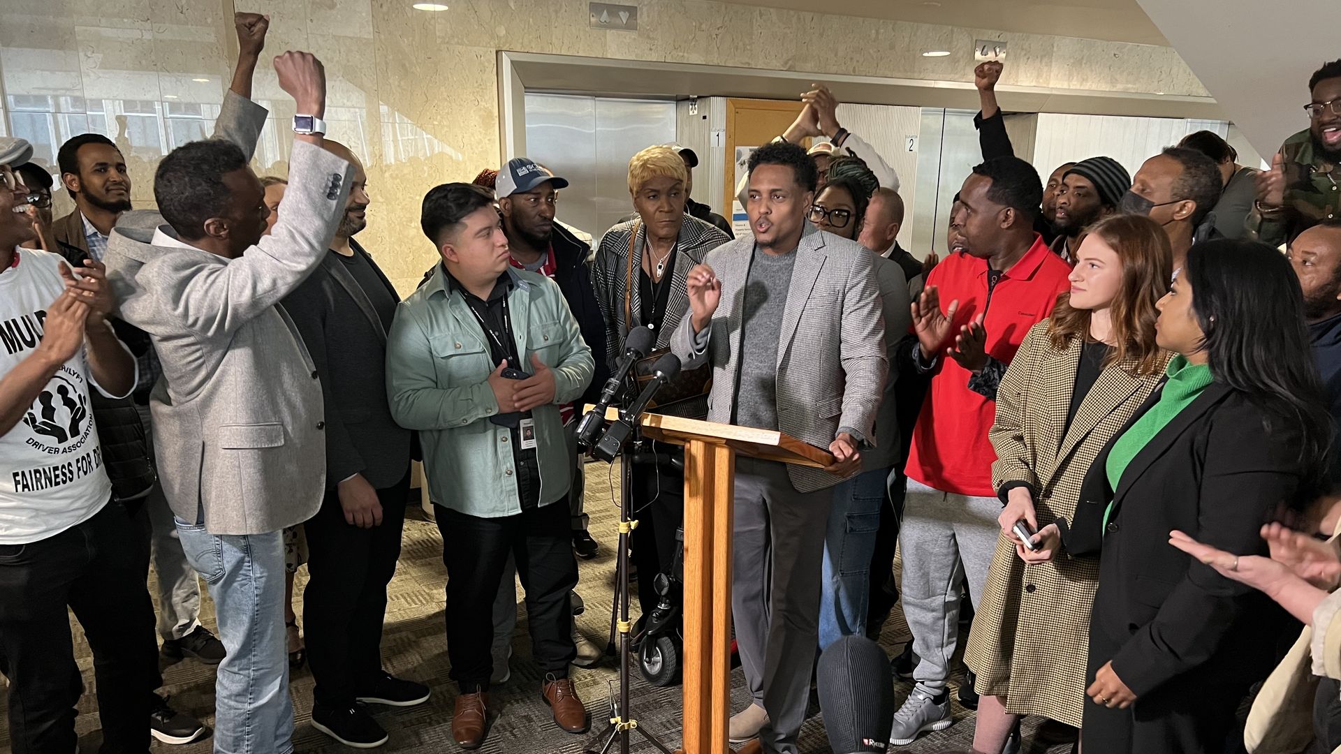 A group of a few men raise their fists in solidarity and celebration as another man speaks at a lectern with a microphone at a press conference.