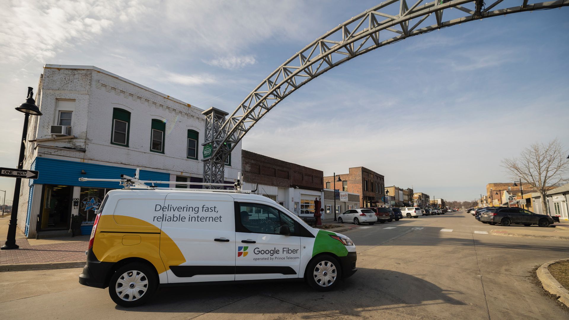 White Google Fiber van with yellow and green accents parked on a small town street under a metal arch, with brick buildings and cars lining the road on a partly cloudy day.