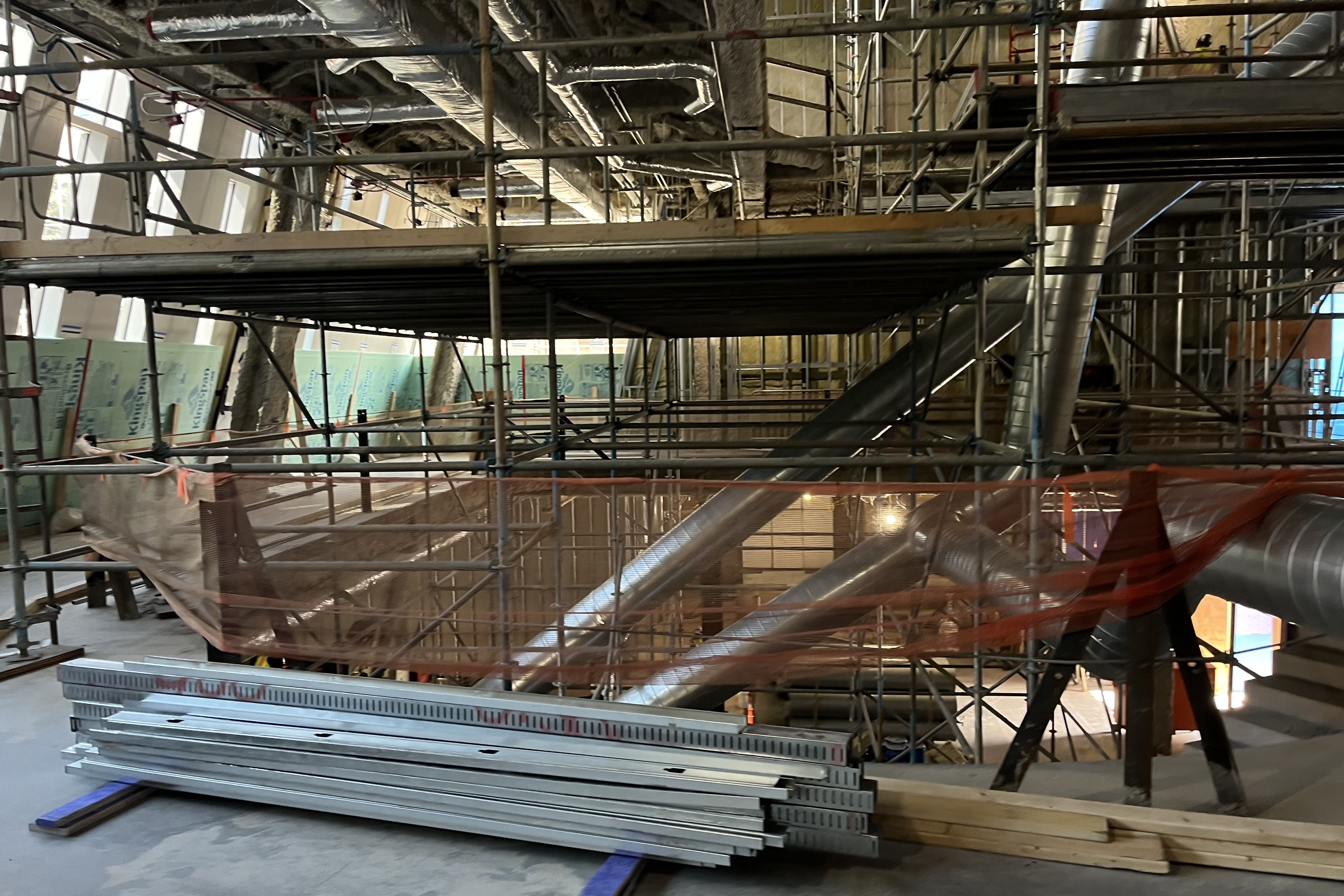 Interior construction site with metal scaffolding, exposed ductwork, insulation paneling, and stacks of metal beams on a concrete floor under natural light from slanted windows.