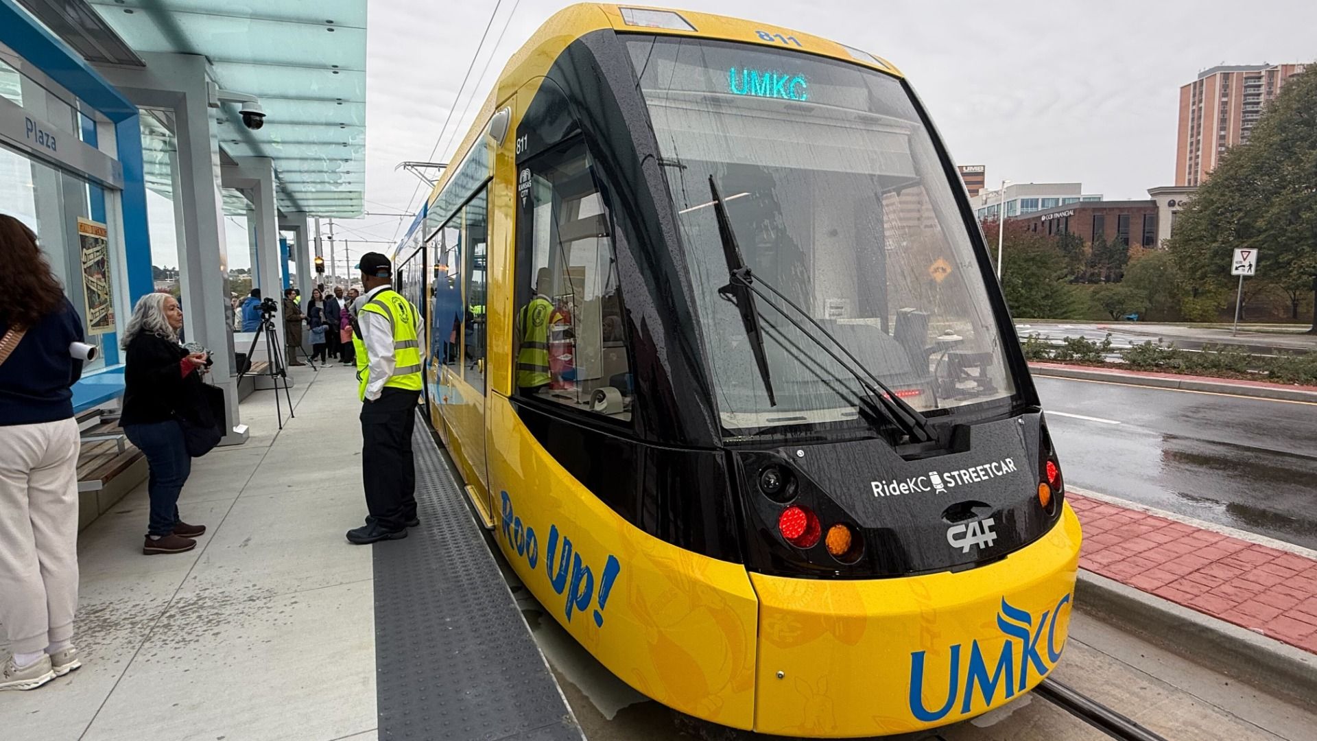 Yellow and black RideKC streetcar at a station platform, displaying UMKC on its sign. People wait nearby on a gray, overcast day with wet pavement.