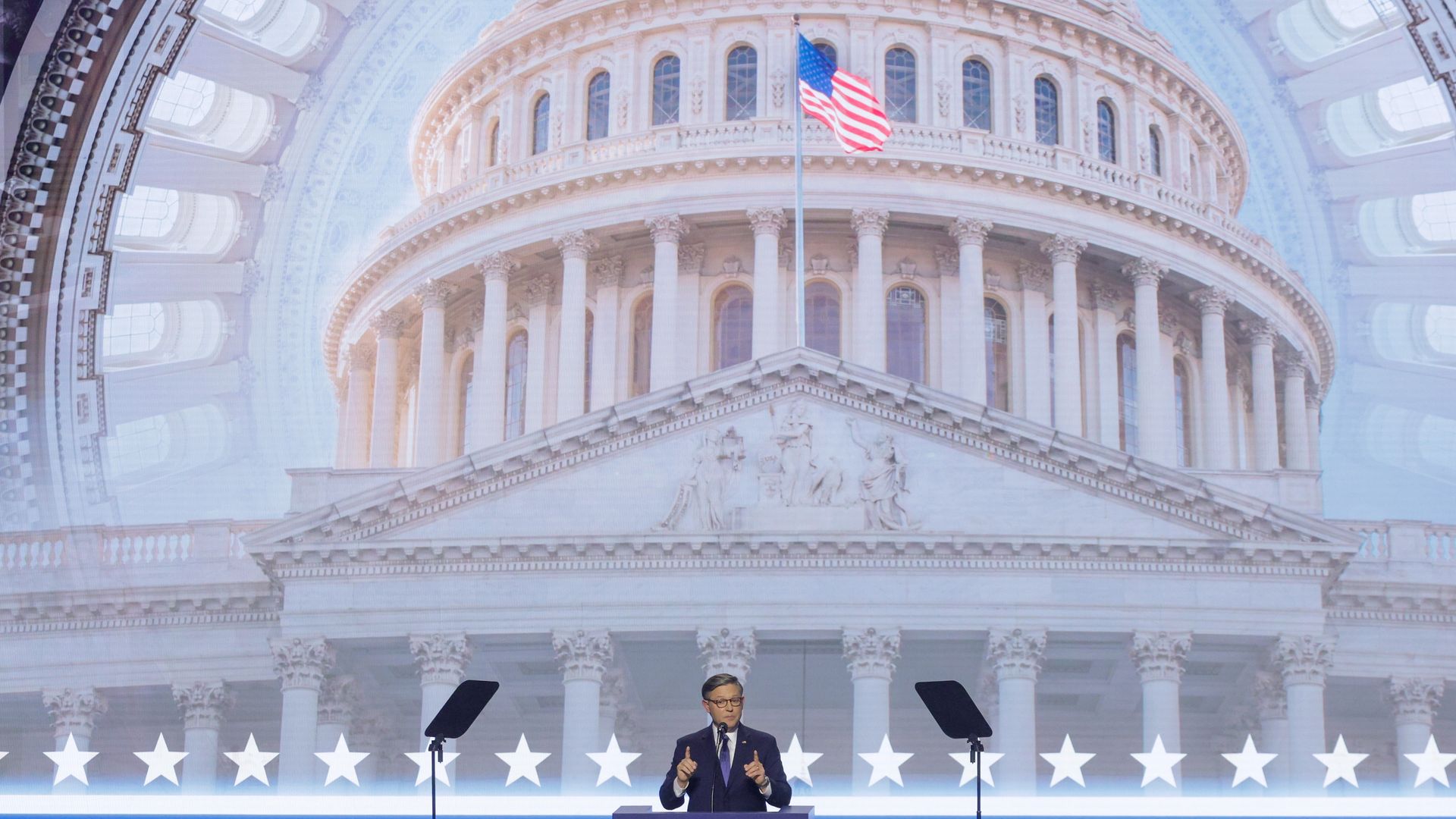 Male speaker in a suit stands at a podium on a blue stage, with a grand domed capitol backdrop and an American flag; white stars line the front edge of the stage.