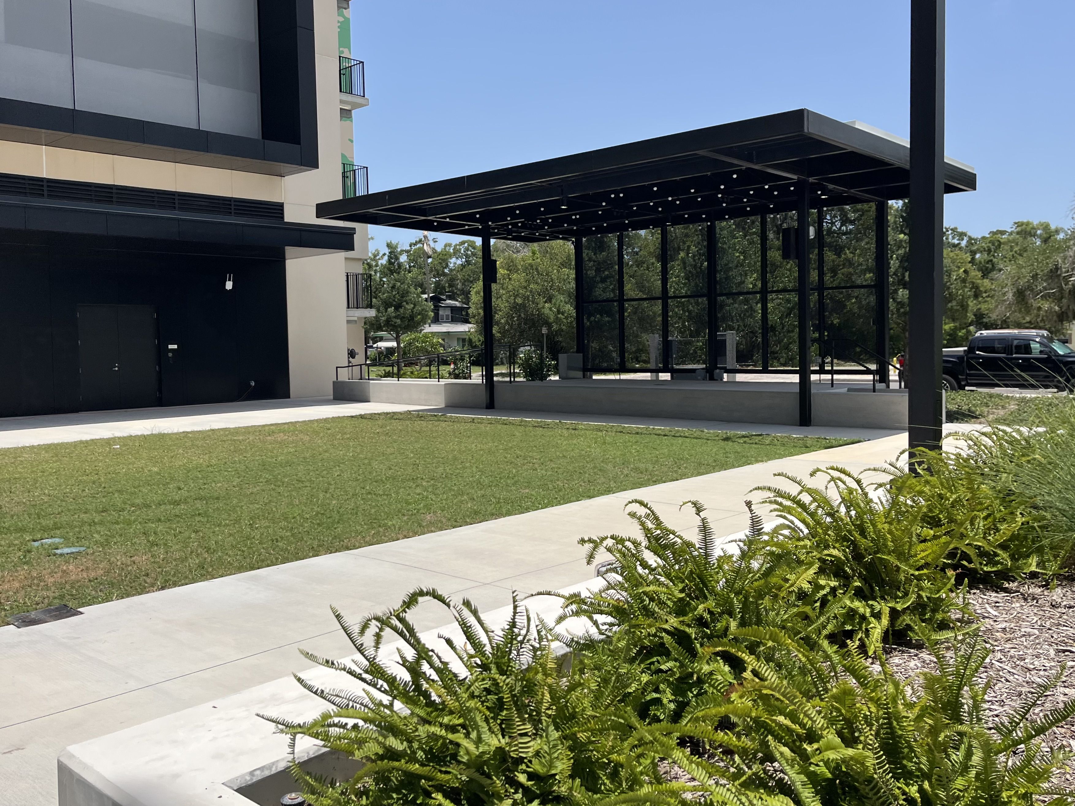 Modern building with dark gray and tan facade. A black metal pavilion with string lights covers a glass-walled area. Green lawn and ferns in the foreground, blue sky, and distant trees with a parked car on the right.