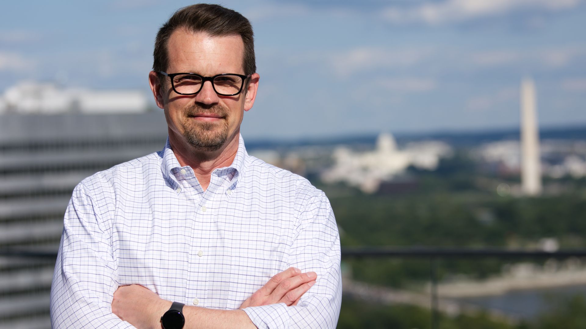 Jason Brown, a man, stands on a balcony overlooking Washington. He's in a white shirt and wearing glasses. A black watch is around his wrist.