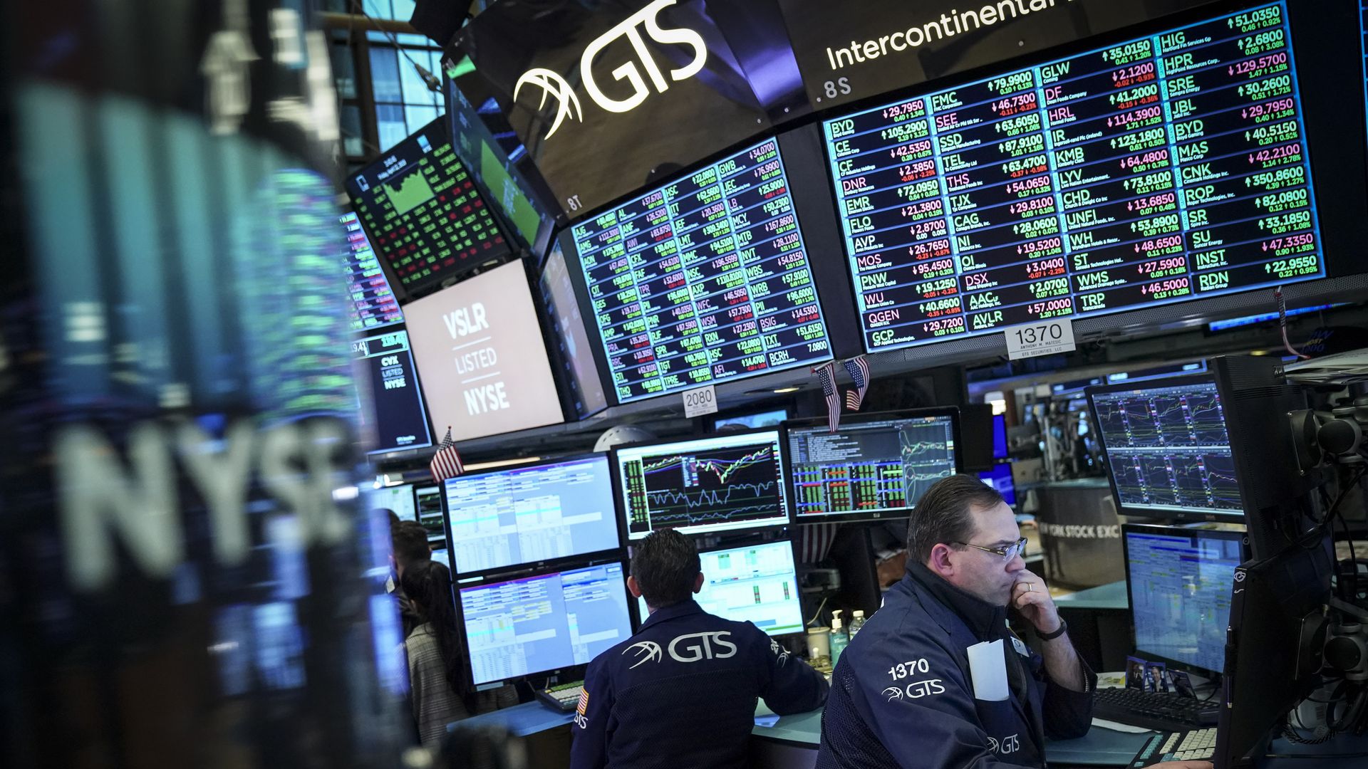 In this image, two men sit at computers underneath large trading board screens at the new york stock exchange.