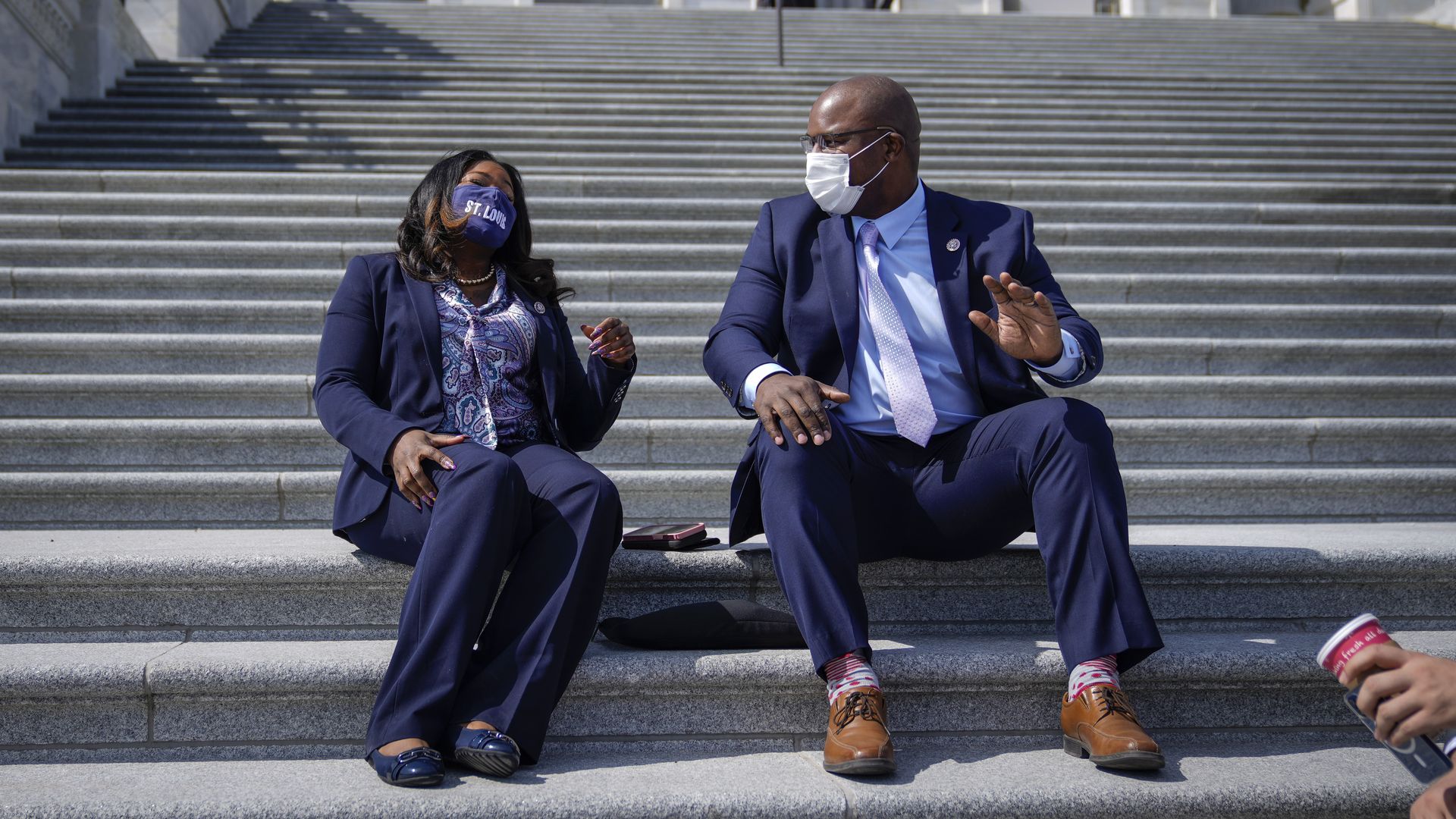 Reps. Cori Bush, wearing a blue pant suit, blue, purple and white blouse and blue "St. Louis" face mask, sits on the Capitol steps with Rep. Jamaal Bowman, wearing a blue suit, light blue shirt, pale purple tie and white face mask.