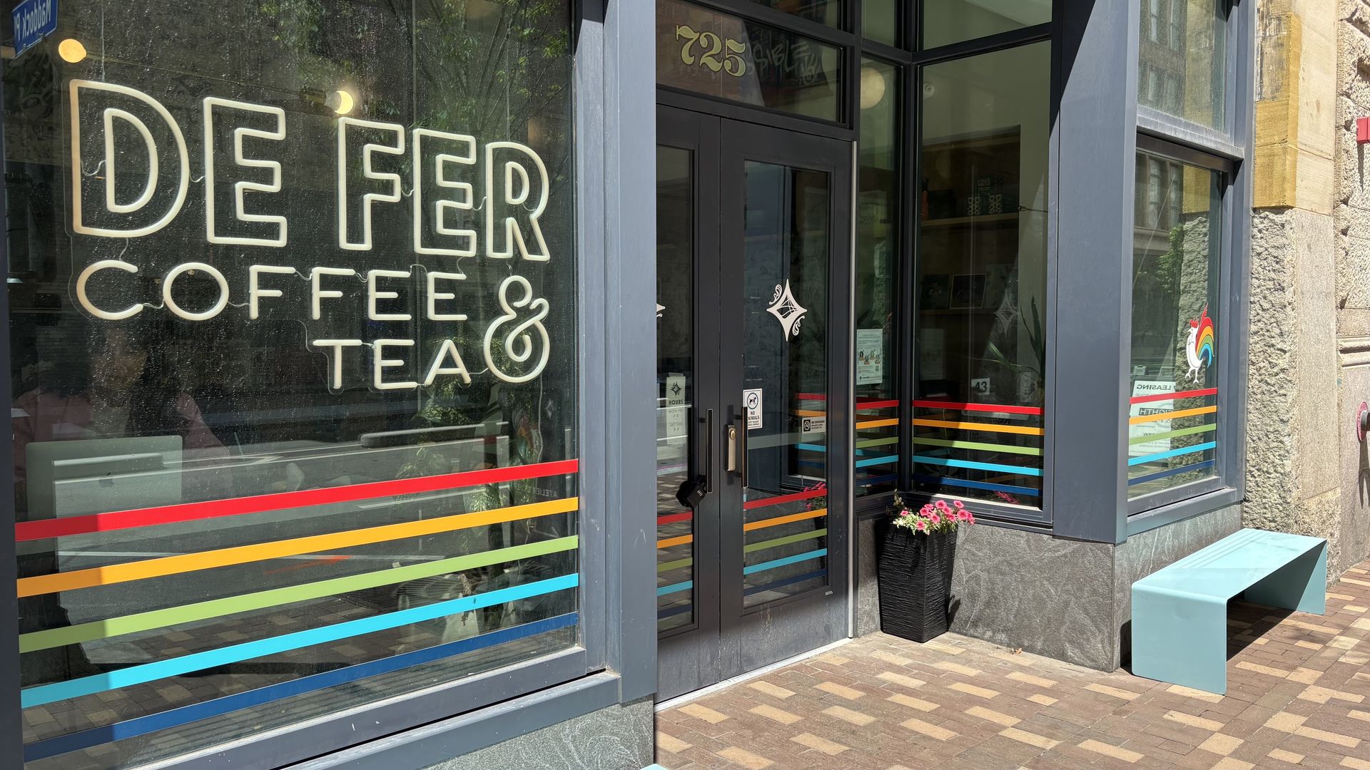 Shopfront with large neon sign "DEEPER COFFEE & TEA" in the left window; rainbow stripe decals on glass, rainbow rooster logo on the right, and a blue bench with flowers outside.