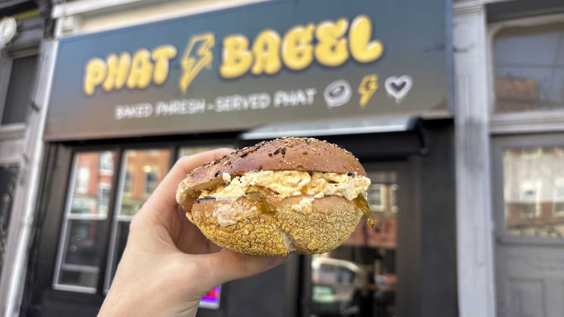 Hand holding an everything bagel sandwich with scrambled eggs and toppings in front of a shop sign reading "PHAT BAGEL" in yellow letters.