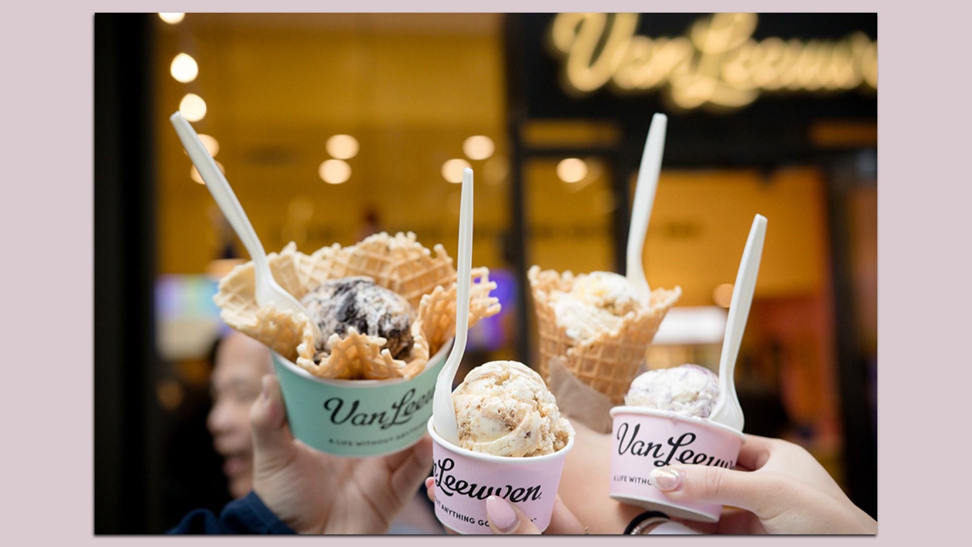Several hands raise pastel ice cream cups with waffle cones and white spoons, showing the Van Leeuwen logo; warm bokeh lights blur in a shop background.