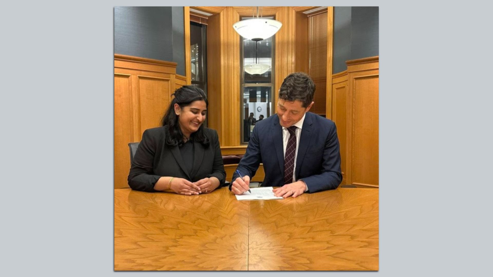 A man in a dark suit signs a document at a wooden table while a woman in black smiles beside him in a wood-paneled room with windows and a hanging light above.