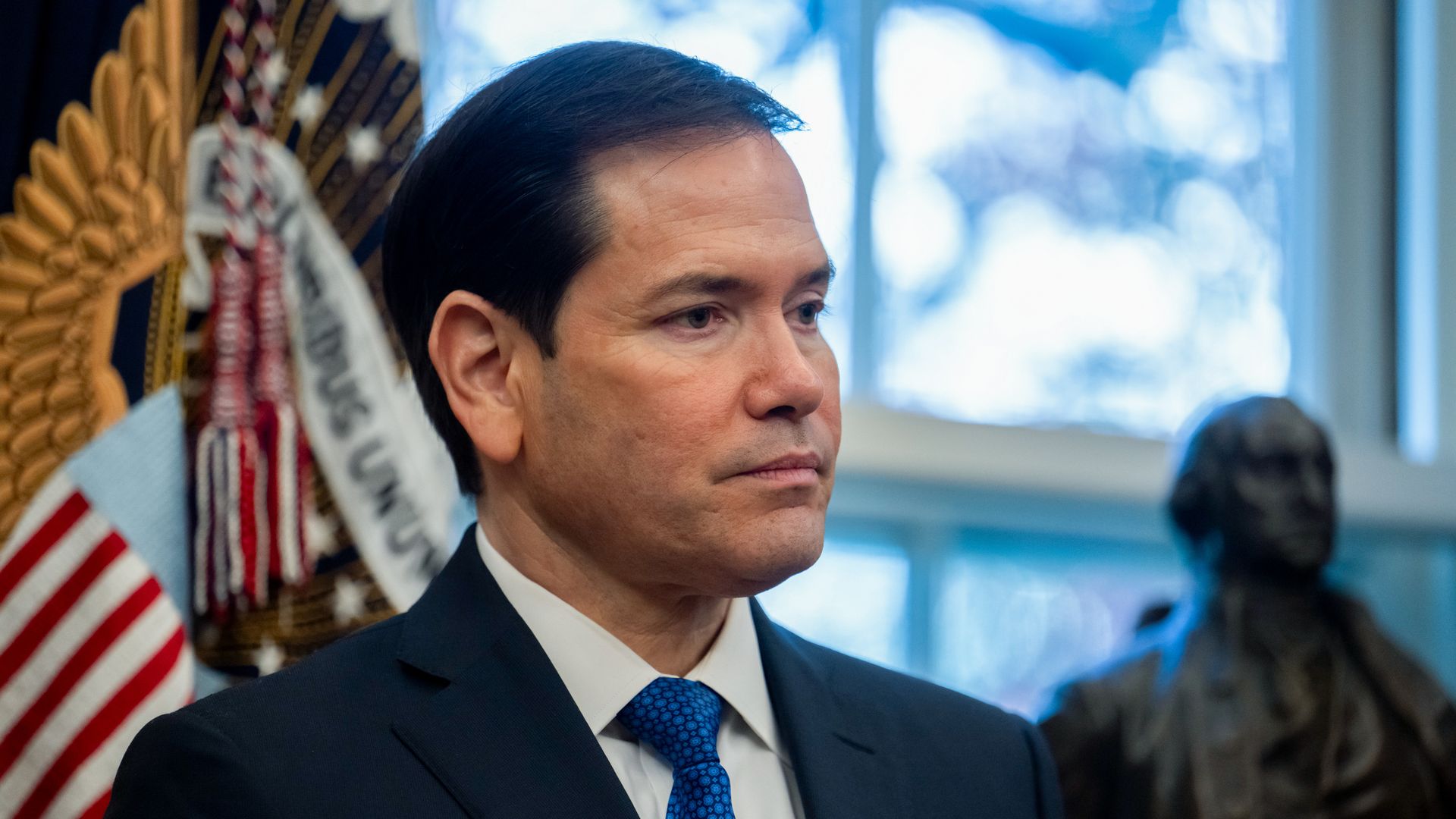 Marco Rubio — wearing a dark suit, a white collared shirt and a blue tie — looks on in the Oval Office.