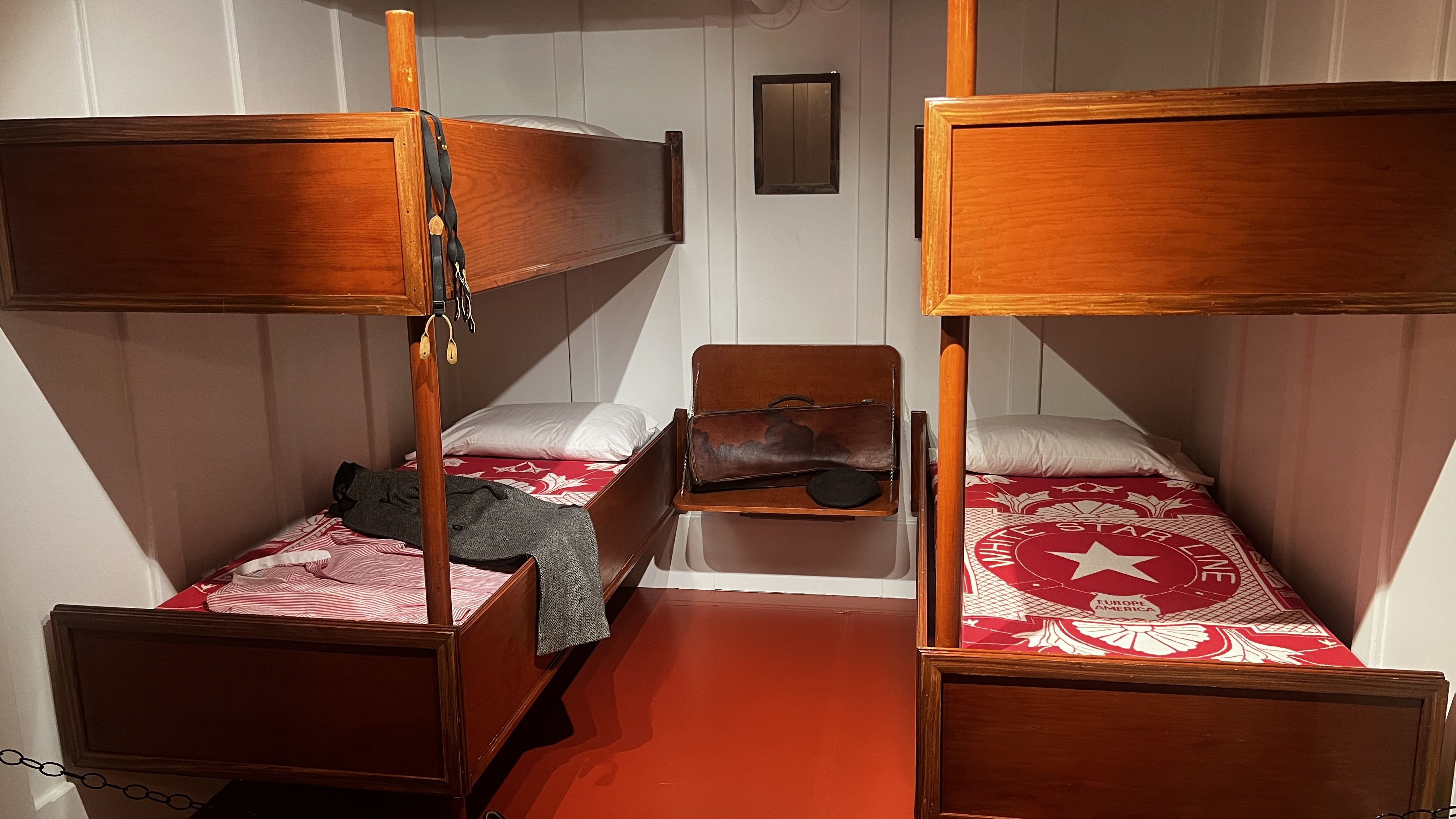 Inside a ship's bunk cabin, two rows of wooden bunks face each other. Right bed has red and white star-pattern sheets; left bed has a gray sweater. A leather bag and a black hat sit on a central shelf.