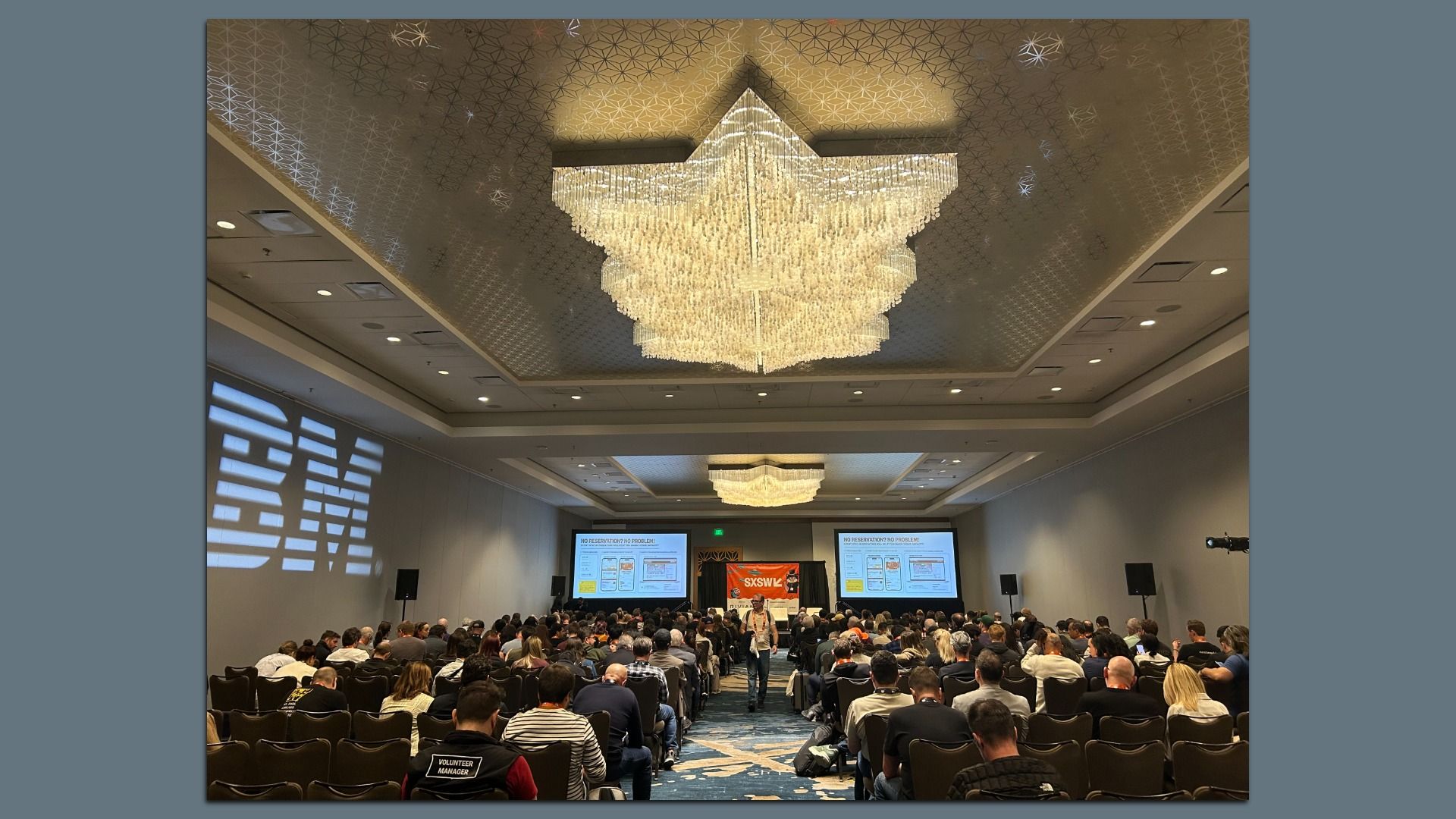 Conference hall with a large crystal chandelier overhead over a crowded audience. Attendees face a stage with two projection screens and an SXSW banner; a person stands in the center aisle.