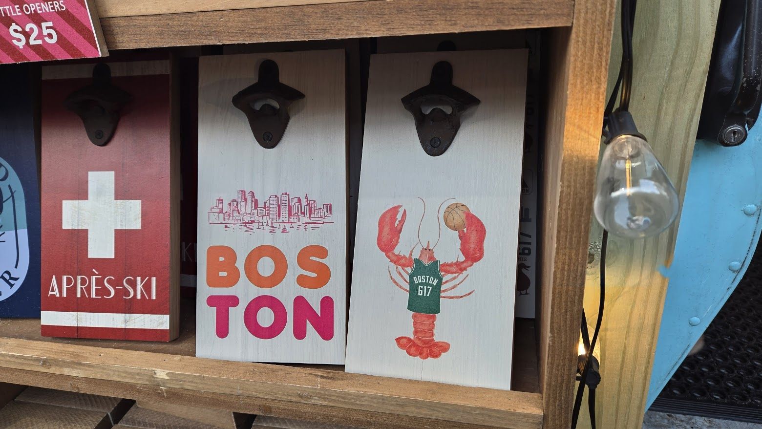 Three wooden bottle openers with metal tops on a shelf. One red with white cross and "APRÈS-SKI", one white with pink and orange "BOSTON" and city skyline, one white with red lobster wearing a green "BOSTON 617" jersey holding a basketball.